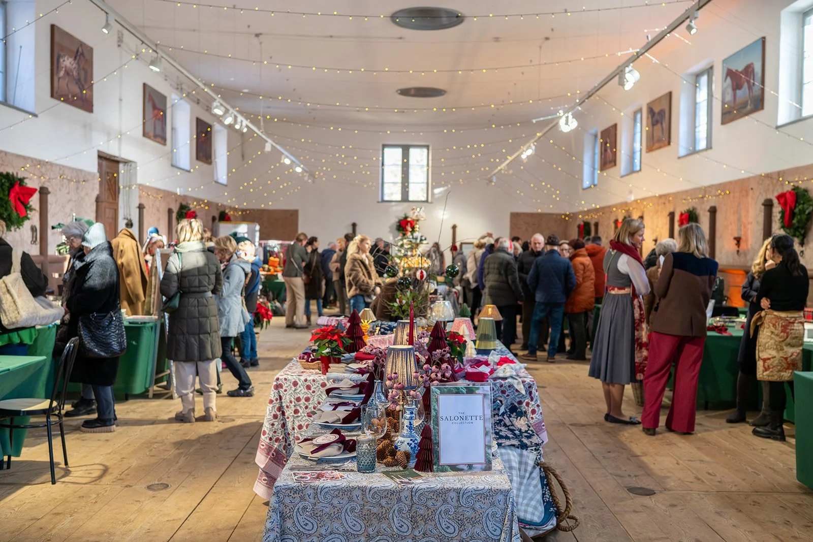 Weihnachtsmarkt in einer großen festlich geschmückten Halle mit vielen Menschen, Tischen mit Dekorationen, Weihnachtsbaum, Lichterketten und Wandbildern mit Pferden.