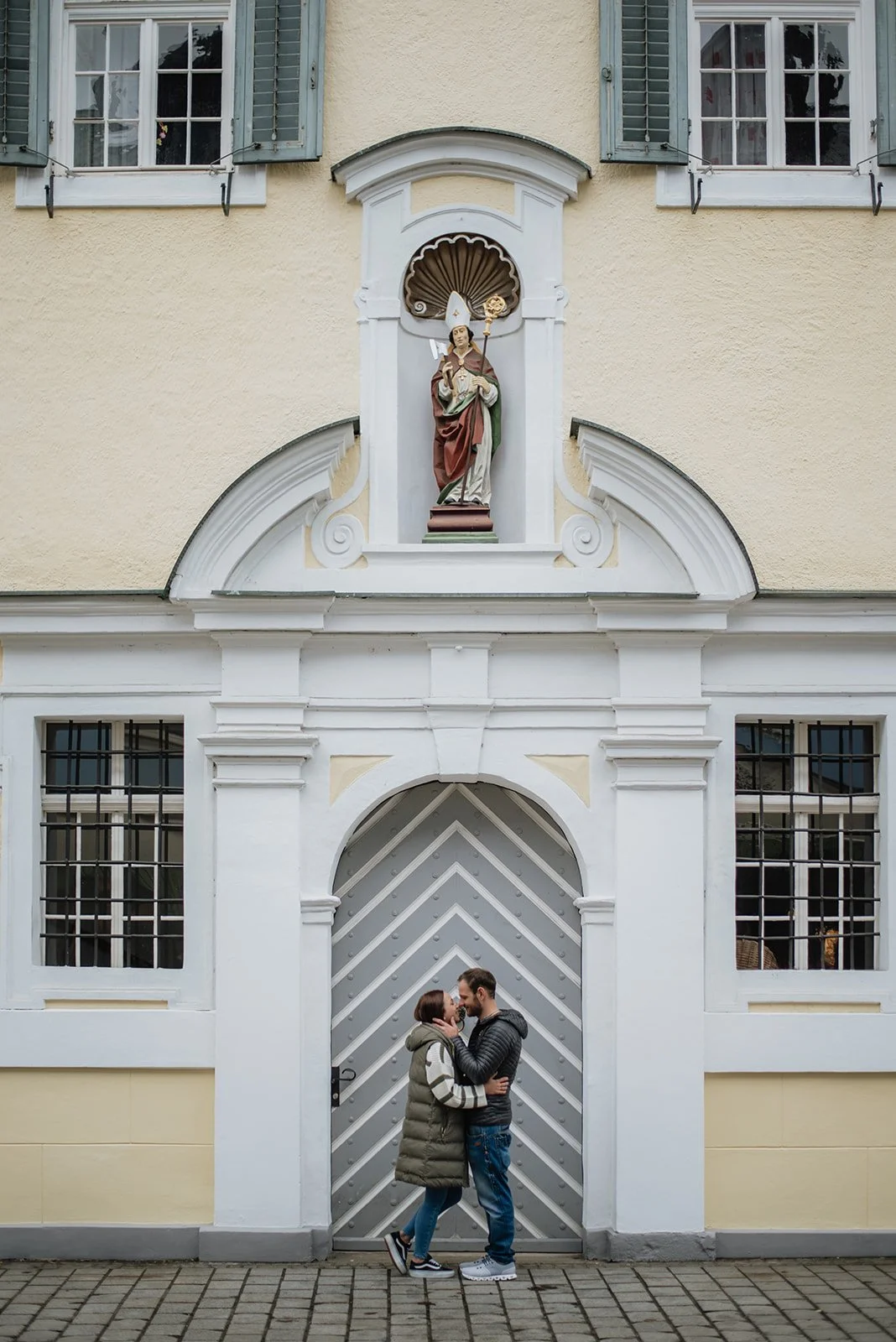 Ein Paar steht vor einem grauen verschlossenen Portal einer Kirche oder eines Gebäudes, das von weißen Fassaden umgeben ist. Über der Tür befindet sich eine Statue eines katholischen Heiligen, vermutlich des heiligen Nikolaus, in einer Nische.