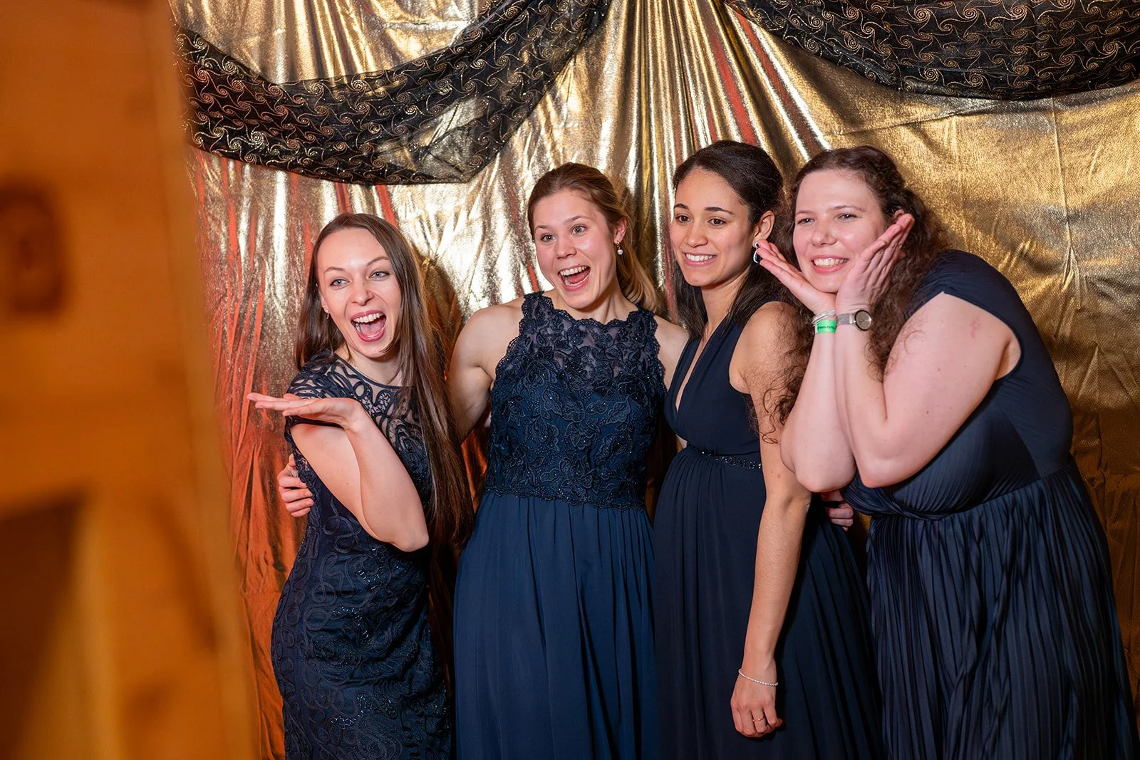 Vier lachende Frauen in dunklen Abendkleidern beim Fotoshooting vor goldener Kulisse.