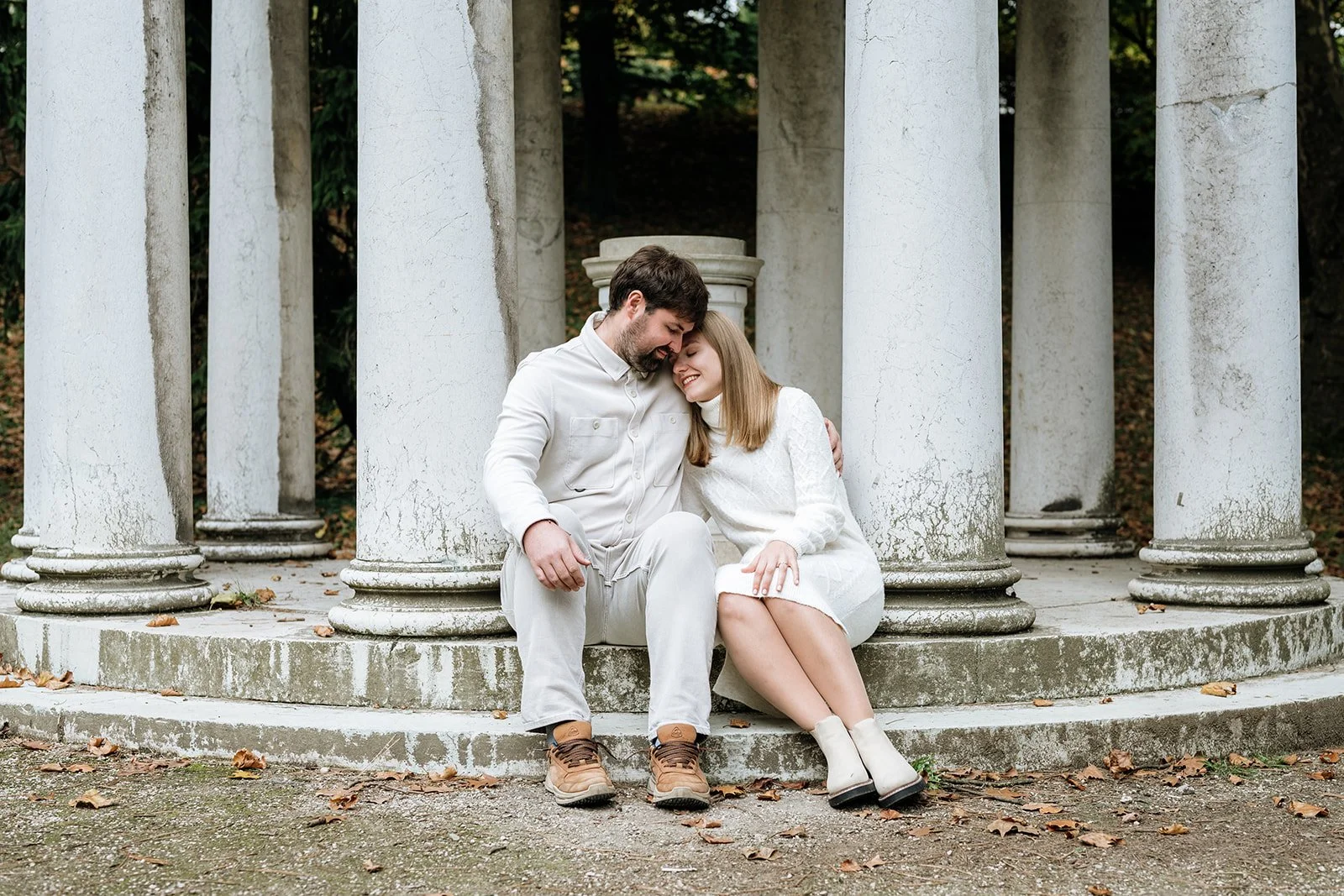 Ein verliebtes Paar sitzt auf den Stufen eines altertümlichen Säulengangs, lächelt und berührt die Stirnen aneinander, umgeben von einem herbstlichen Ambiente.