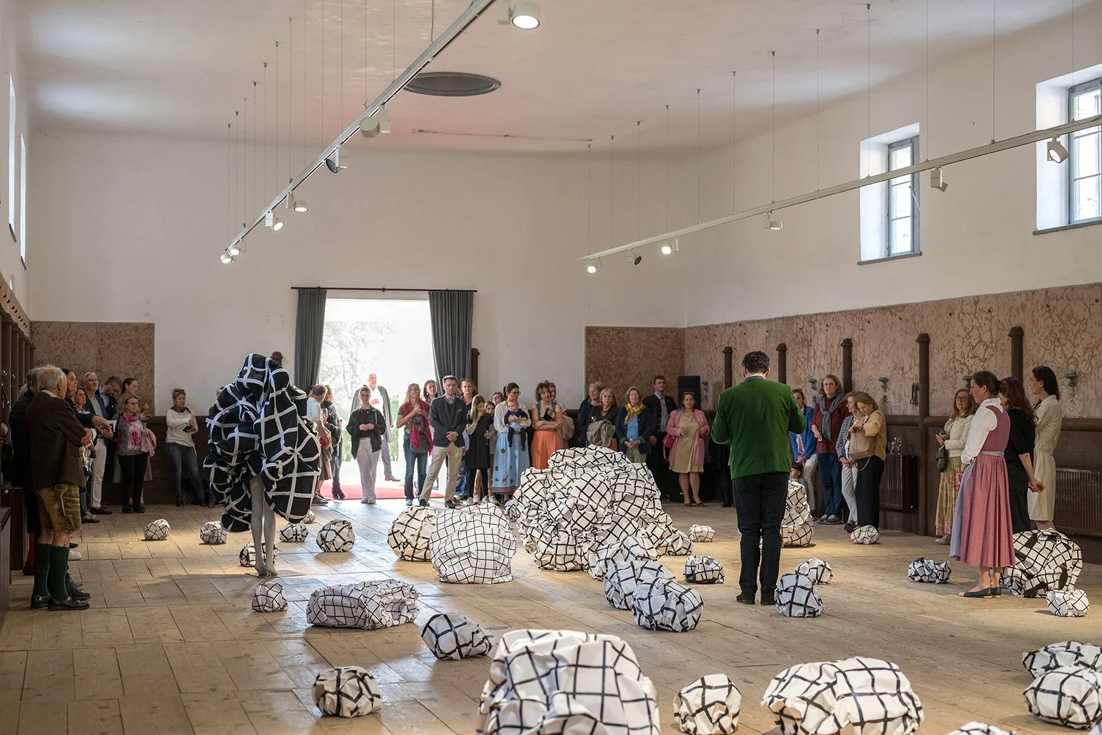 Eine Gruppe von Menschen beobachtet eine Kunstausstellung in einem Raum mit Holzboden und weißen Wänden. Bei den Kunstwerken handelt es sich um große und kleine Papierkugeln, die mit schwarzen und weißen Linien bemalt sind.