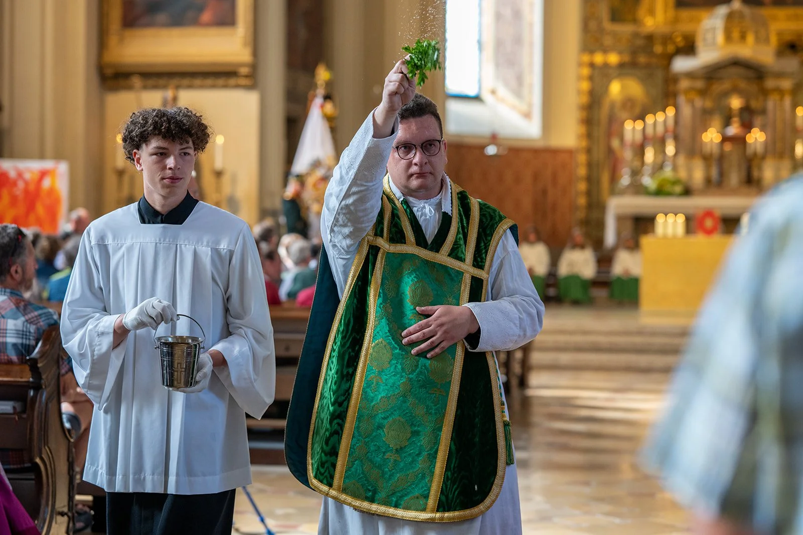 Ein Priester in einer Kirche gießt Wasser über die Hand einer Person während eines religiösen Rituals an einem Altar, im Hintergrund befinden sich mehrere Gläubige.