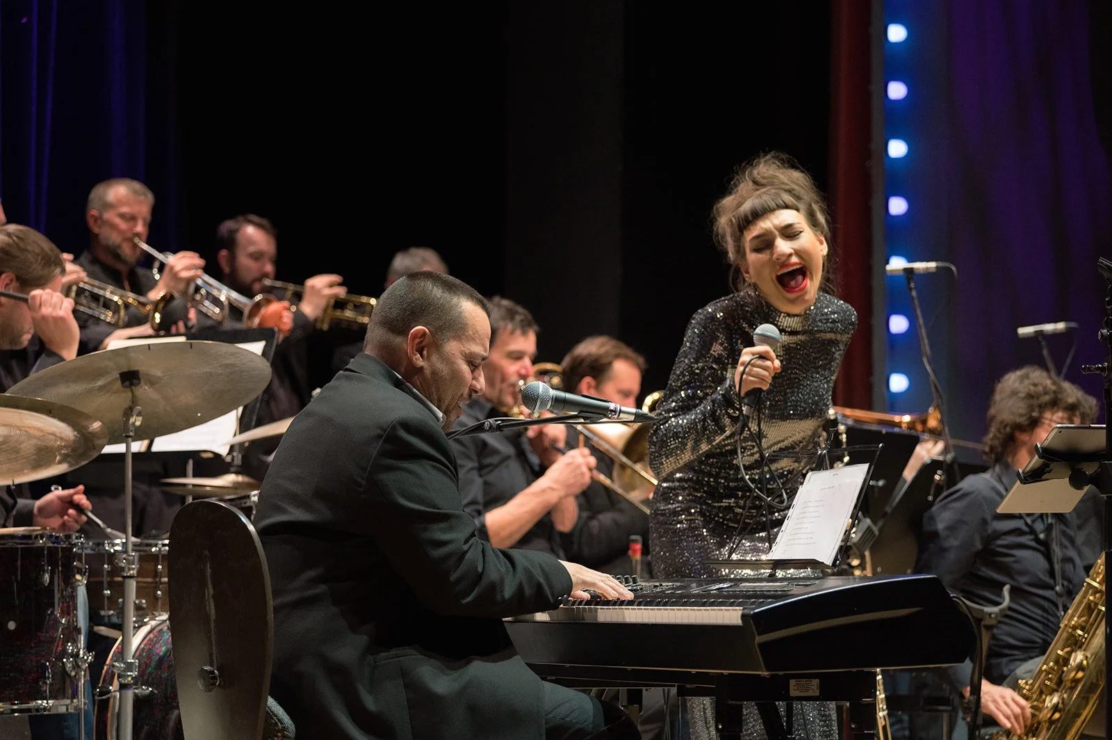 Eine Jazzband mit einem weiblichen Sänger, die auf einer Bühne performen, begleitet von einem Pianisten und einem Blasorchester im Hintergrund.