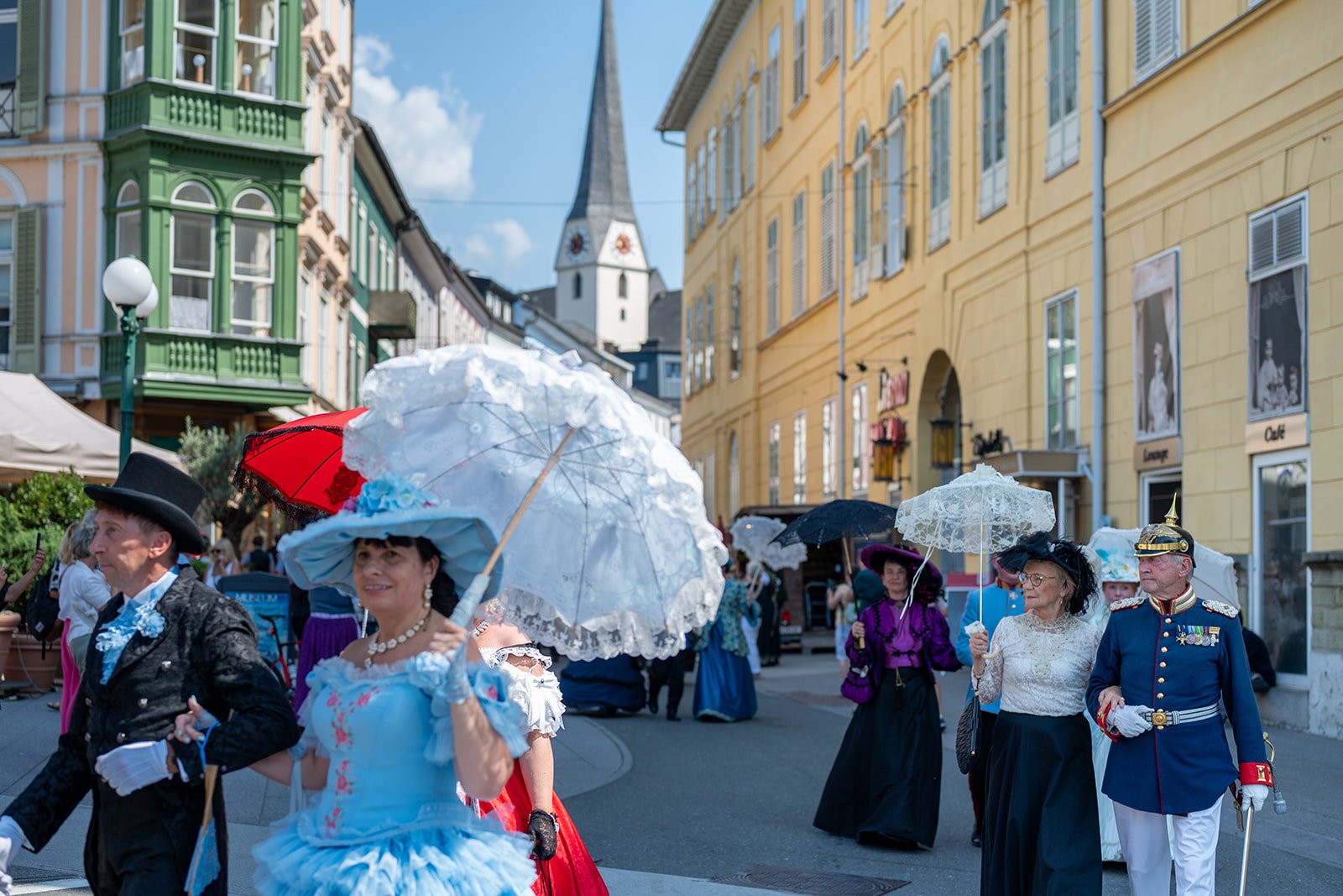 Menschen in historischen Kostümen bei einem Festumzug auf einer Straße in der Stadt, mit bunten Gebäuden im Hintergrund.
