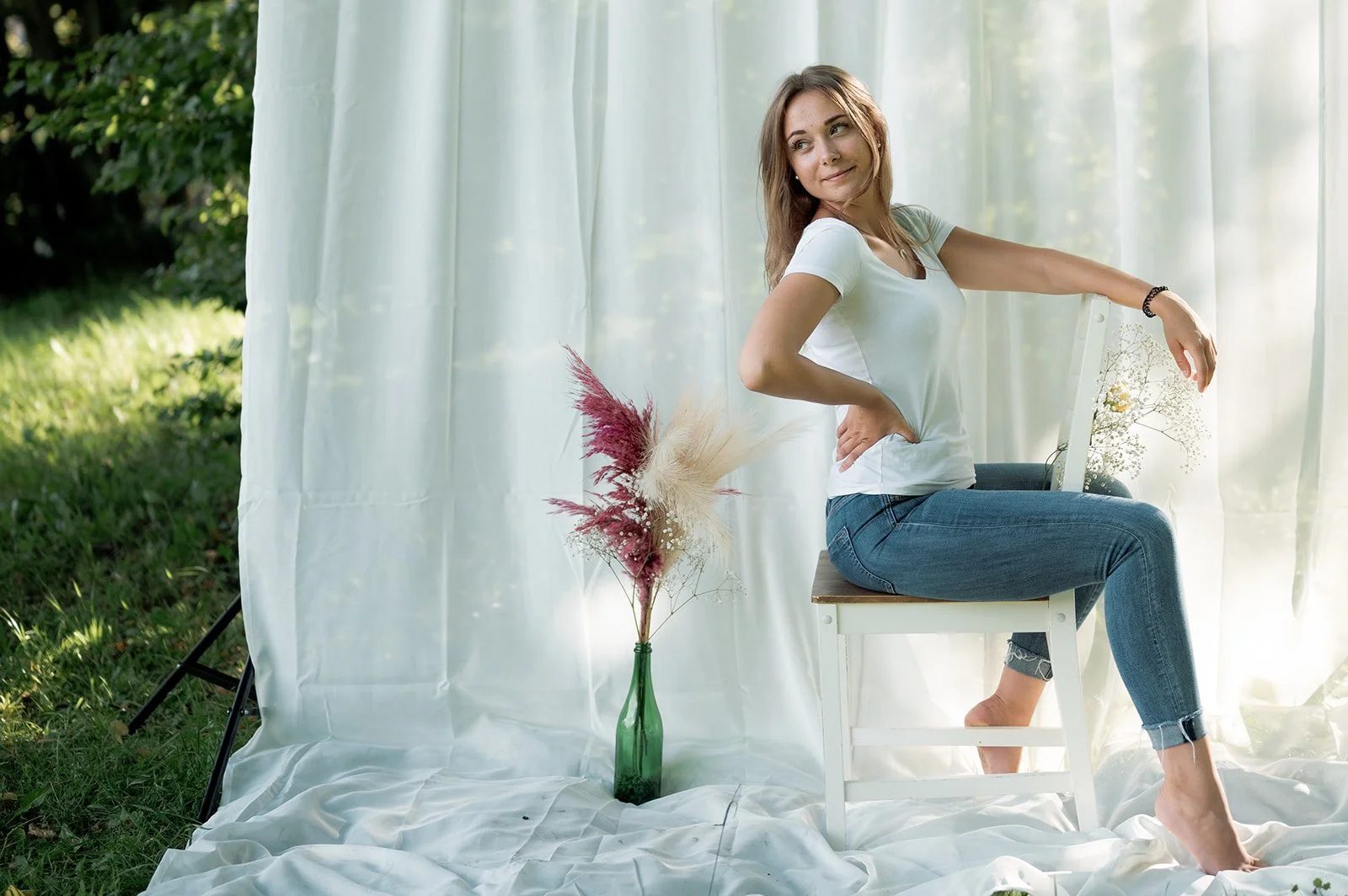 Junge Frau sitzt auf einem Stuhl mit weißen Stoff als Hintergrund, draußen auf dem Gras, mit einer Vase voller getrockneter Blumen neben ihr.
