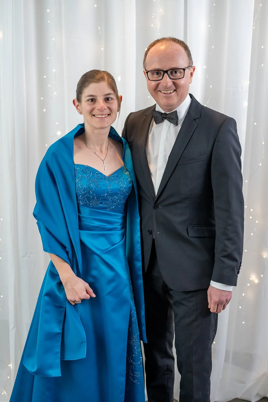 Ein Mann und eine Frau stehen nebeneinander vor einem weißen Hintergrund mit Lichterketten. Sie tragen formelle Kleidung, die Frau in einem blauen Kleid und der Mann in einem schwarzen Anzug mit Fliege.