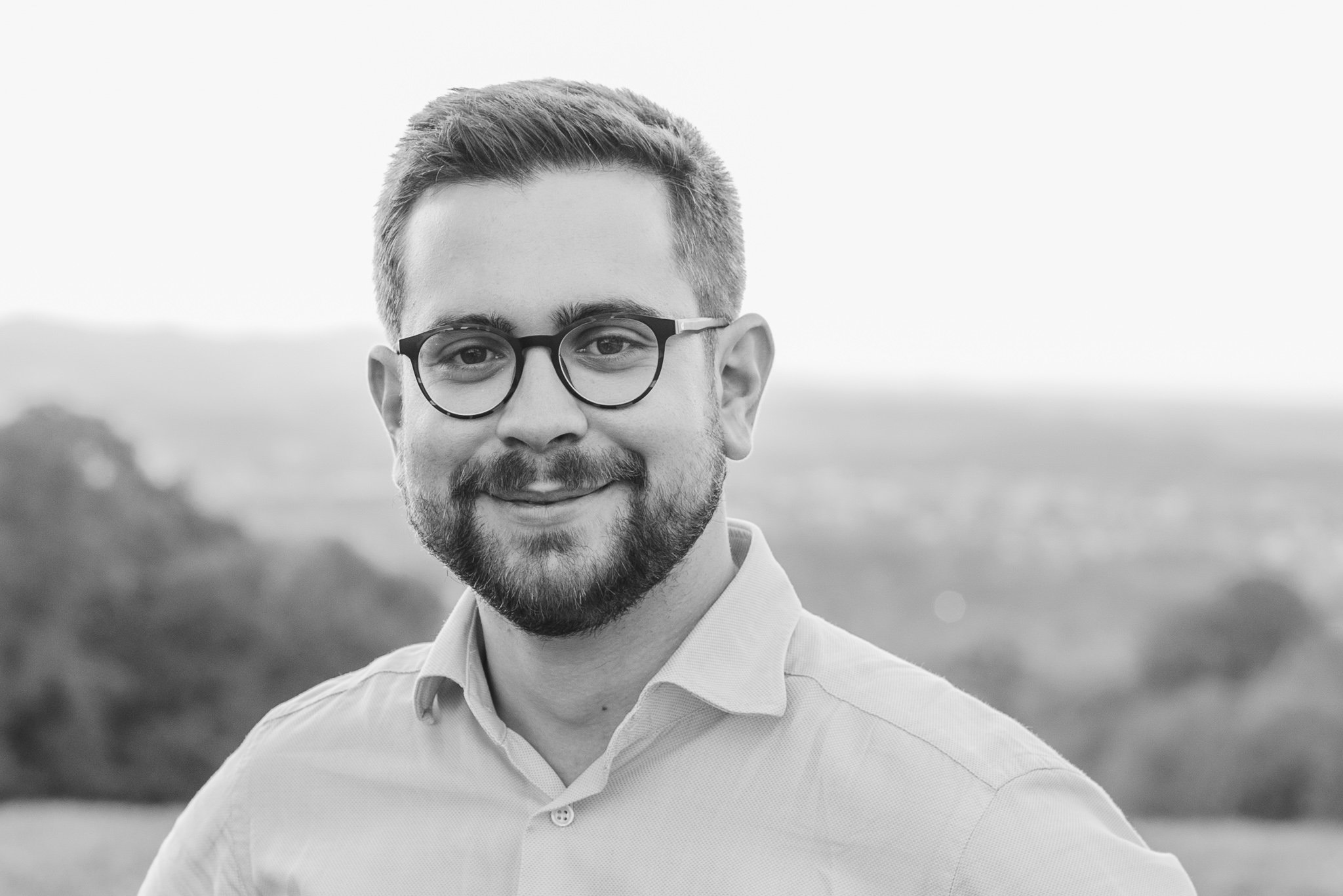 Schwarz-Weiß-Foto eines lächelnden Mannes mit Brille und Bart, trägt ein Hemd, im Hintergrund eine ländliche Landschaft