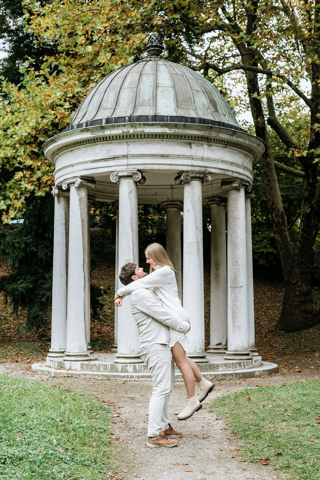 Ein Mann hebt eine Frau vor einer Pavillon im Park. Die Frau lächelt und sieht den Mann an. Es ist herbstlich mit bunten Blättern an den Bäumen im Hintergrund.