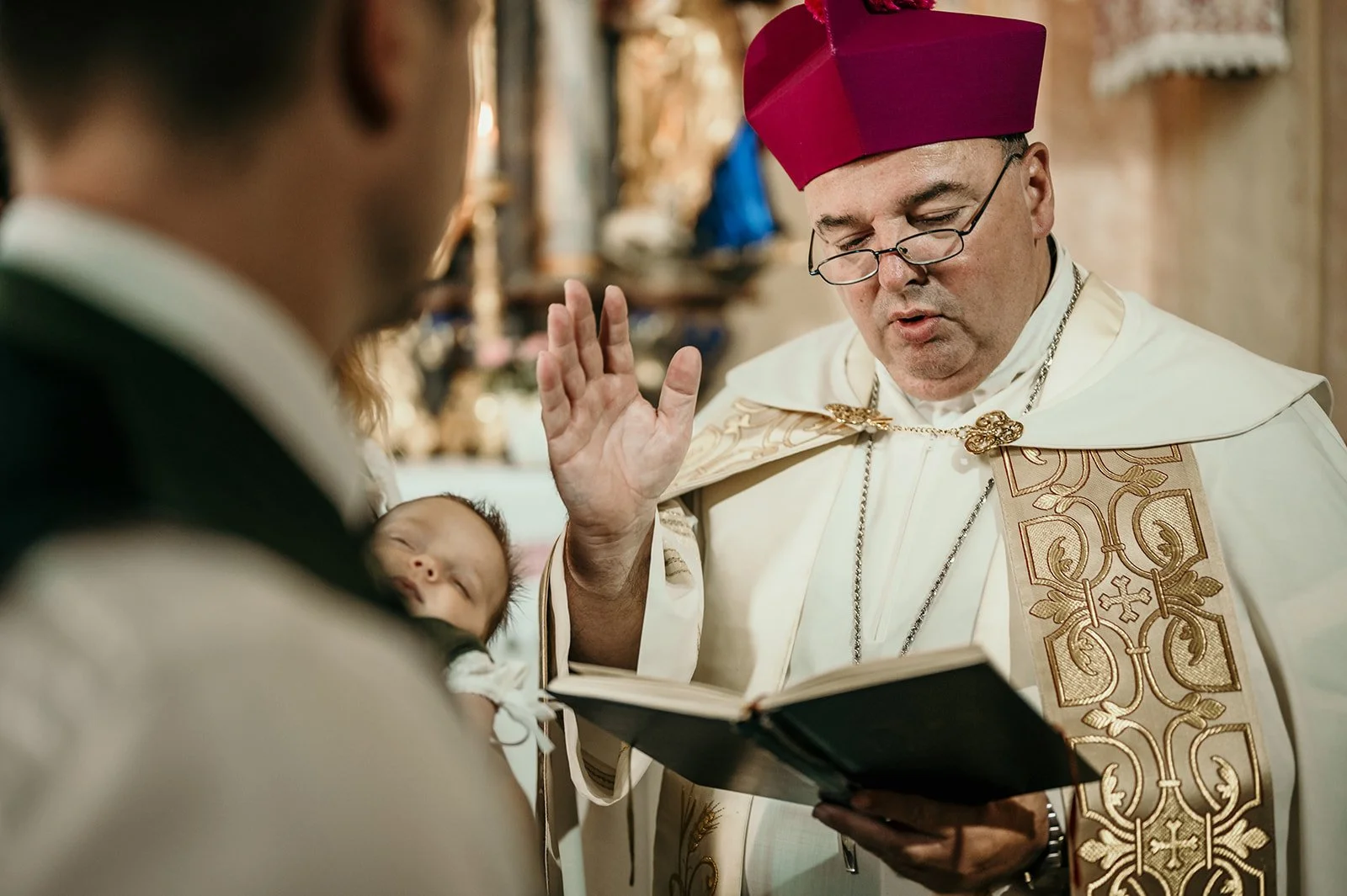 Ein Priester in weißen Gewand mit goldener Verzierung und rotem Hut, der eine Zeremonie in einer Kirche durchführt, während er ein Buch liest und einen Segen erteilt, mit einem schlafenden Baby in den Armen eines Mannes.