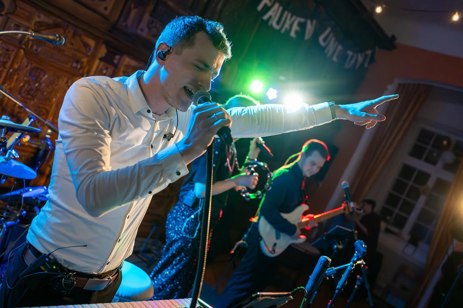 Eine Band live auf einer Bühne, ein Sänger mit Headset, der in ein Mikrofon singt, mit ausgestrecktem Arm, im Hintergrund Musiker mit Instrumenten, bunte Beleuchtung, Holz-Wand, Fenster in einem Raum mit Vorhängen.
