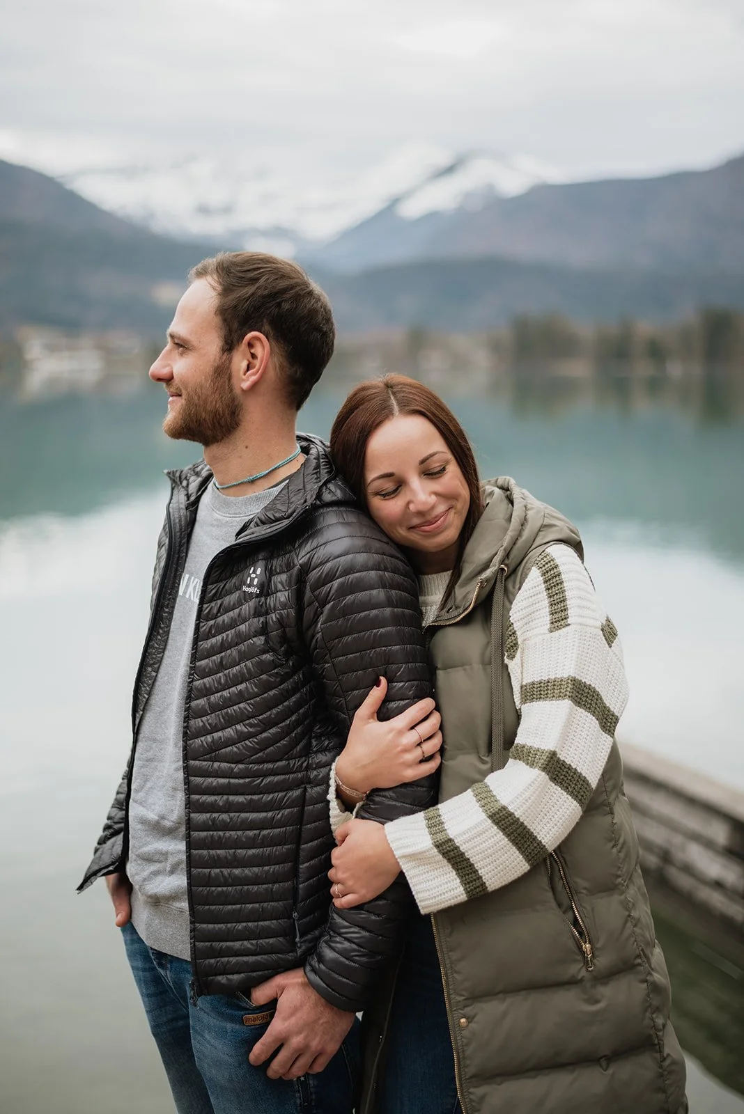 Ein Mann und eine Frau stehen am See in den Bergen, sie umarmen sich und genießen den Moment, im Hintergrund sind schneebedeckte Berge zu sehen.