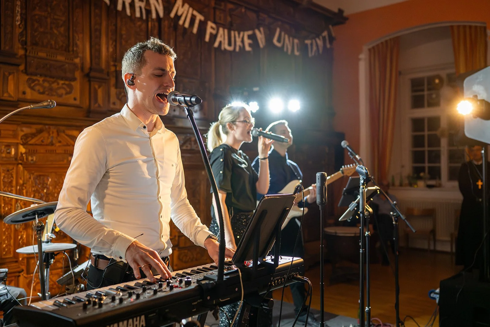 Eine Band mit einem männlichen Keyboardspieler und zwei Sängerinnen im Hintergrund, die in einem festlich dekorierten Raum während eines Konzerts auftreten.
