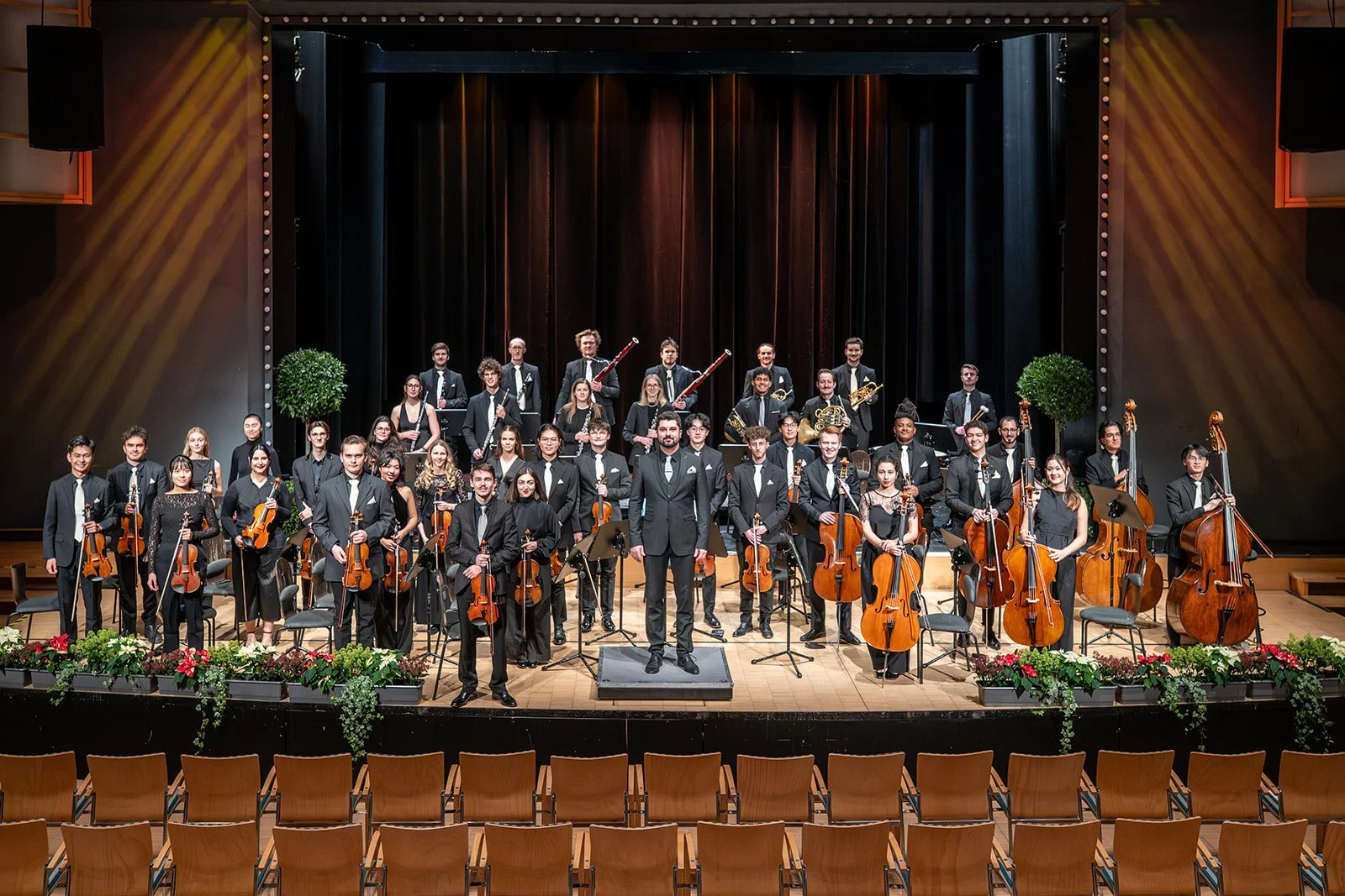 Ein Orchester in schwarzen Anzügen auf einer Bühne mit Holzboden, im Hintergrund ein dunkler Vorhang, und vorne eine Podest mit einem Dirigenten. Es gibt städtisches Dekor mit Pflanzen und roten Blumen vor der Bühne, und leere Stühle im Vordergrund.