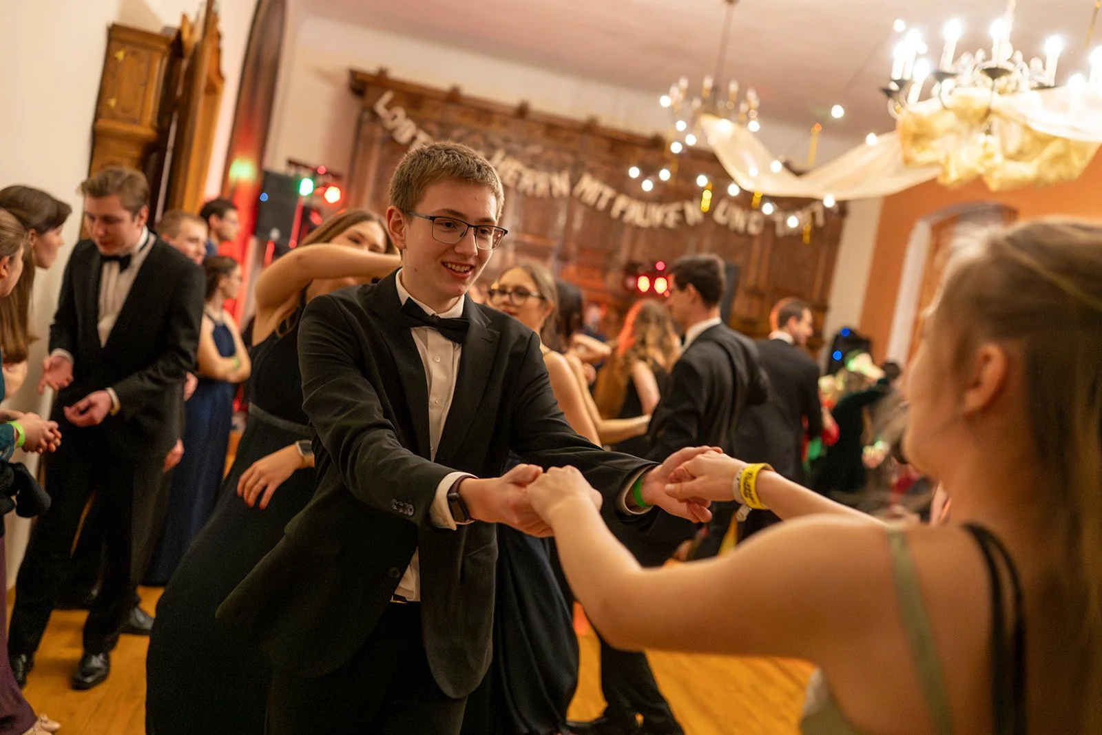 Junge Menschen tanzen auf einer Party in formeller Kleidung, im Hintergrund Dekorationen und Lichter.