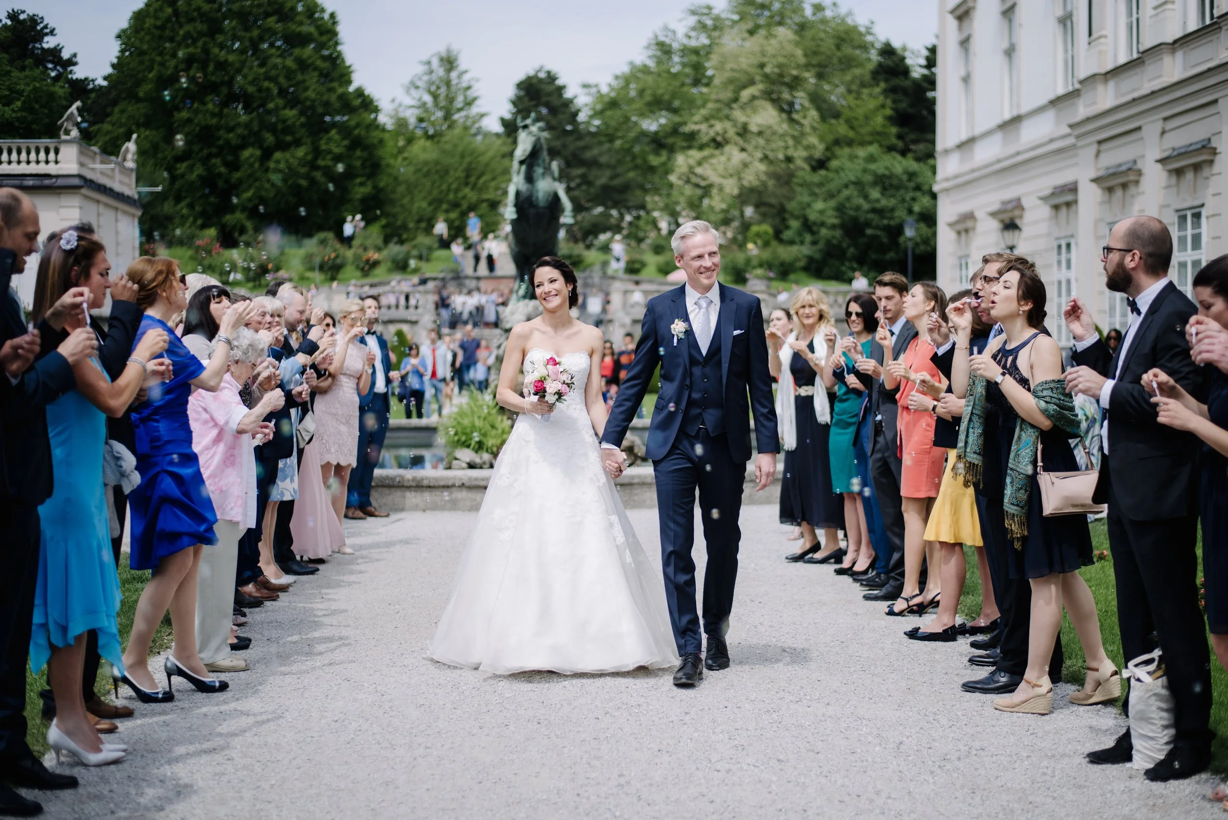 Ein Brautpaar läuft Hand in Hand durch eine Menschenmenge bei einer Hochzeit im Freien, umgeben von Gästen, die Bonbons oder Konfetti werfen, mit einem Brunnen und Bäumen im Hintergrund.