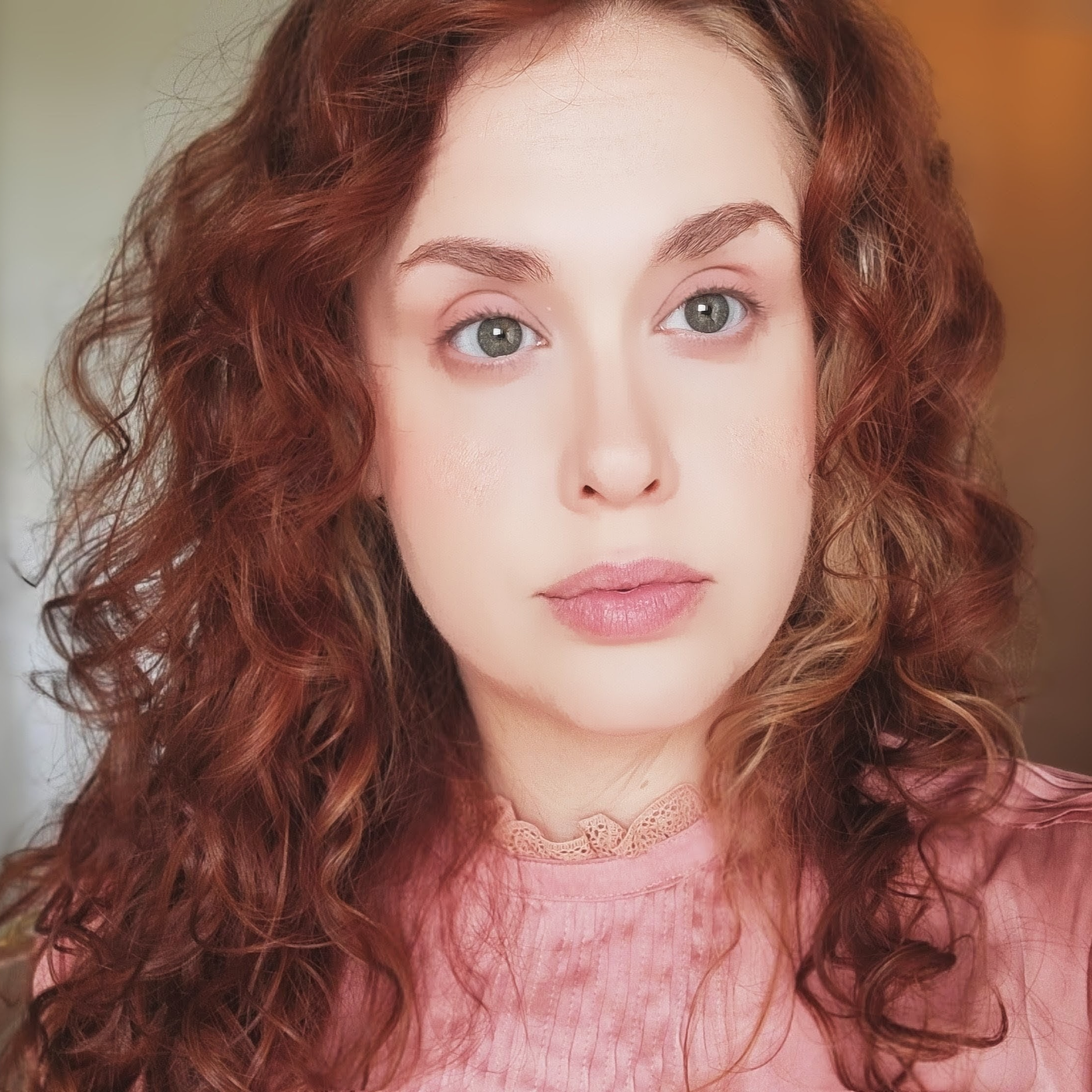 Close-up of a woman with curly red hair wearing a pink top with lace detail, and natural makeup with soft pink lips.