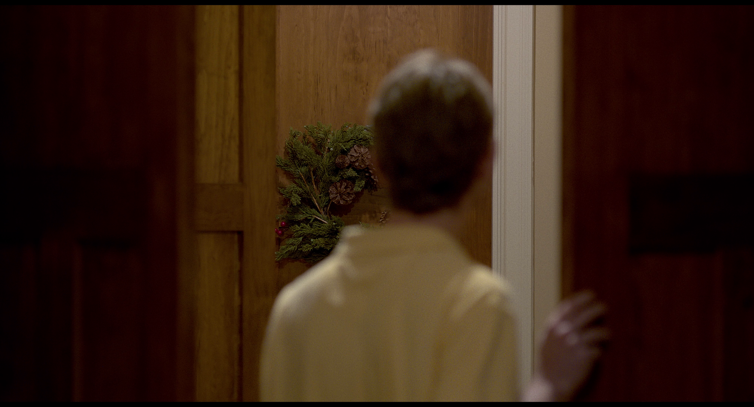 A person with short hair, wearing a light-colored shirt, peeking through a doorway with a holiday wreath on the door.
