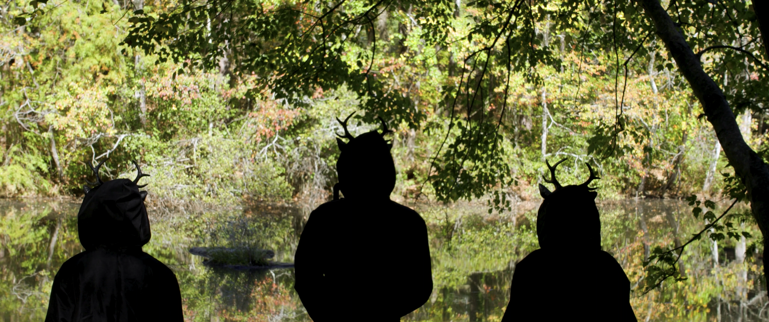 Silhouettes of three children with deer antlers on their heads facing a lake and forest with autumn foliage