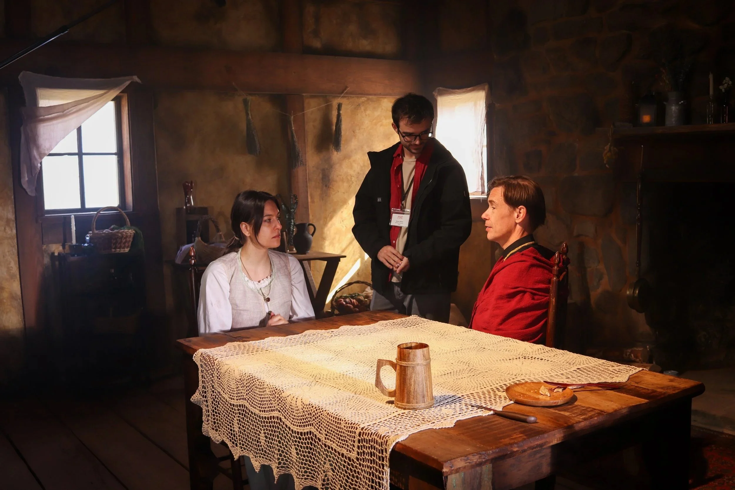 A scene inside a rustic room with stone walls and wooden furniture featuring three people: two seated at a wooden table and one standing. The woman on the left has dark hair and wears a white shirt; the man on the right has light hair and wears a red