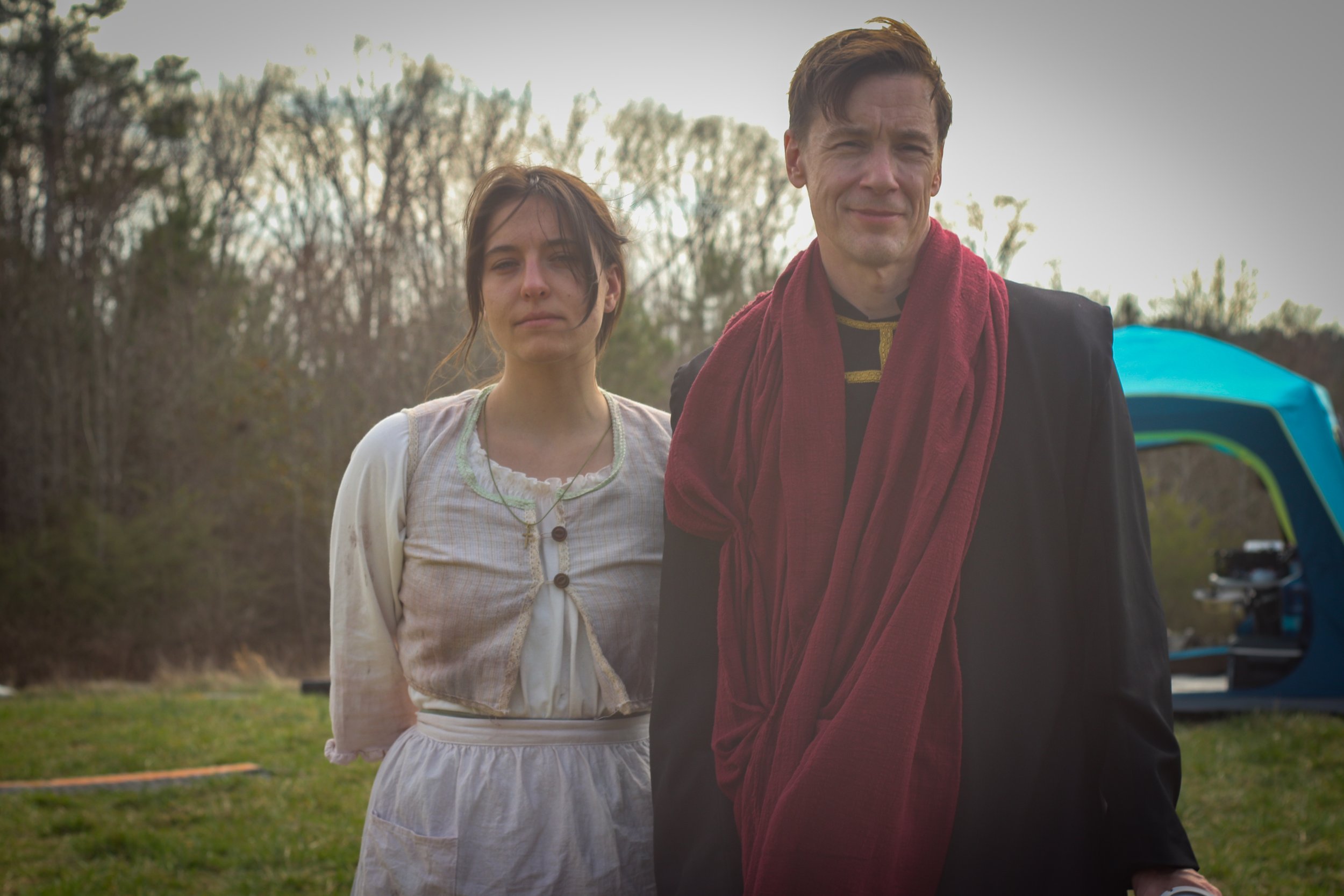 Two people standing outdoors with trees and a blue tent in the background, one man and one woman. The man is wearing a black coat with a red scarf, smiling slightly, and the woman has a neutral expression, wearing a light-colored dress and cardigan.