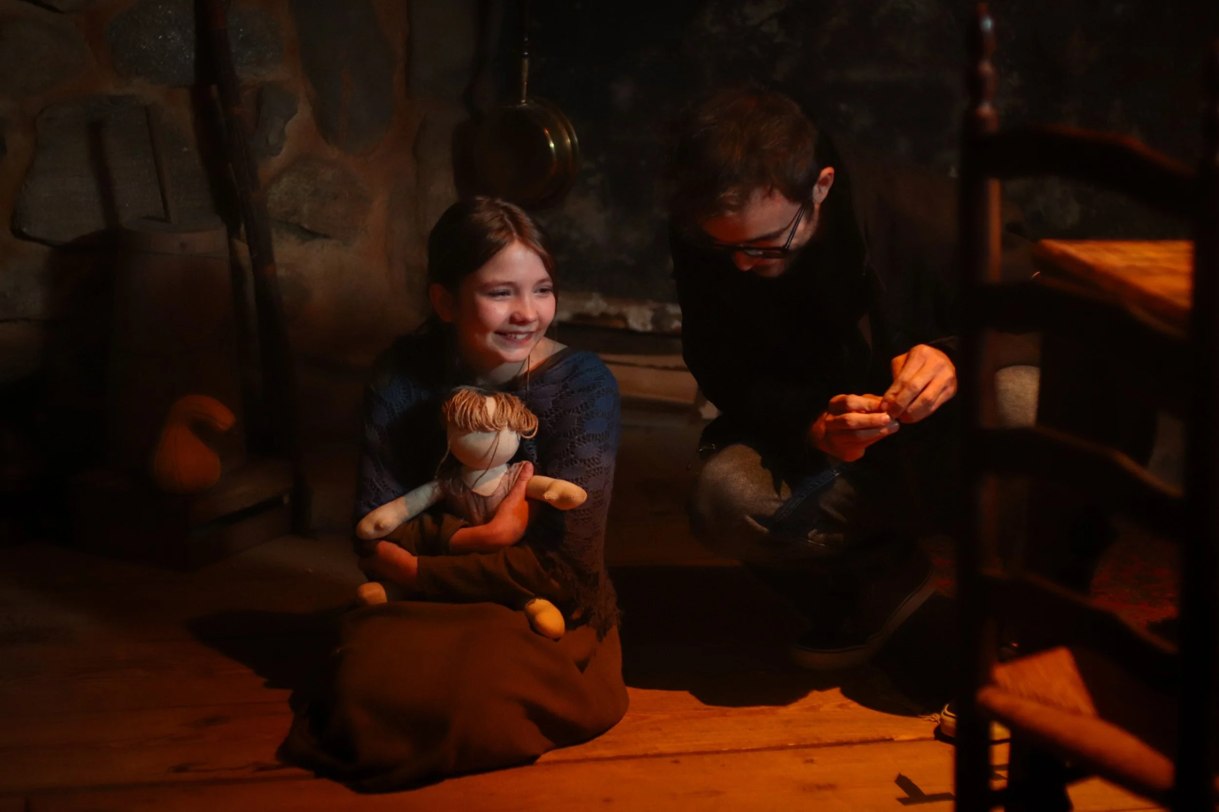 A young girl sitting on the wooden floor of a dimly lit room, smiling and holding a homemade sock puppet, while an adult man kneels next to her, engaging with her, with a stone wall and hanging pots in the background.