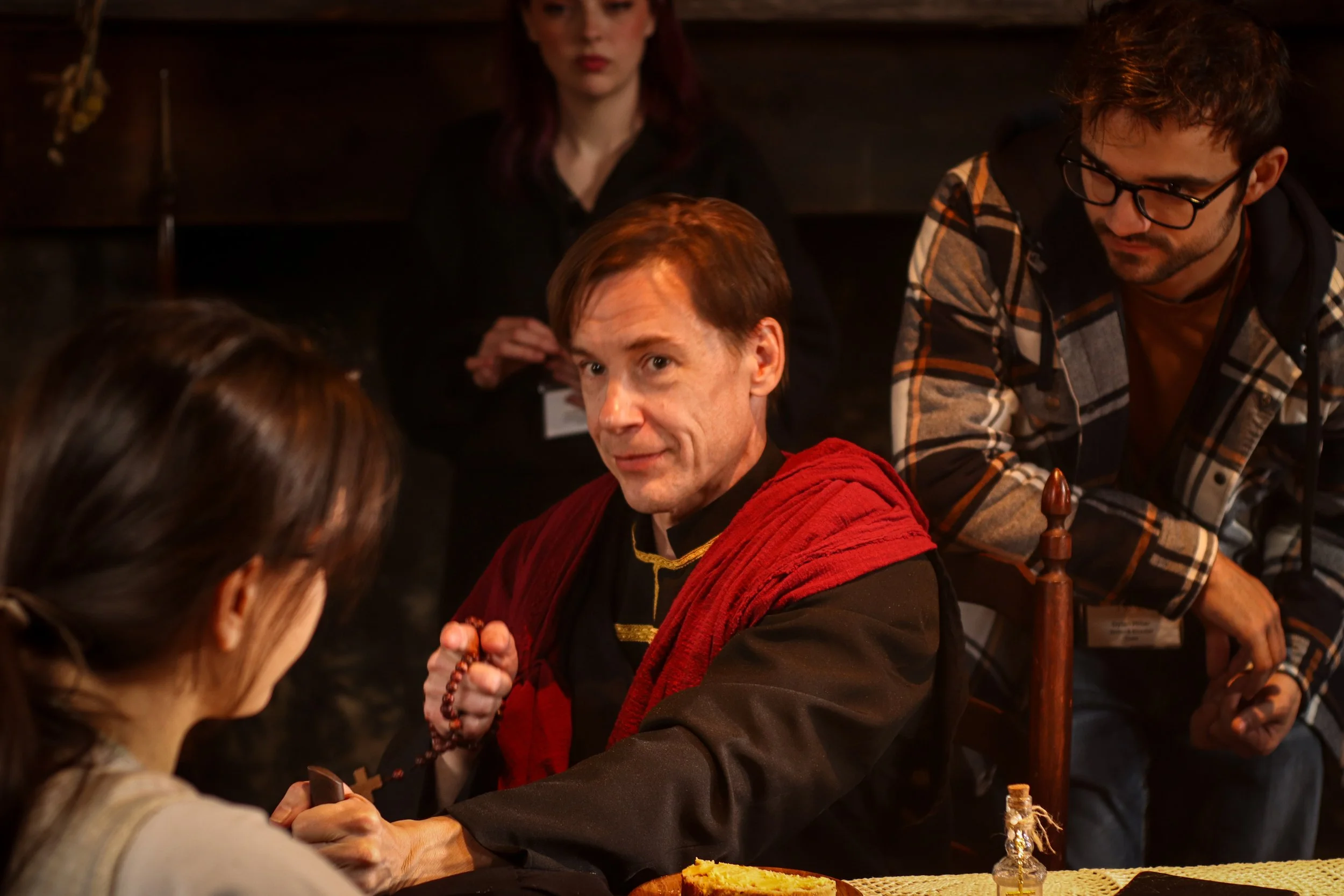 A man dressed as a monk, holding prayer beads, sitting at a table with a woman whose face is not visible, while a young man with glasses stands nearby, and a woman with red hair sits in the background.
