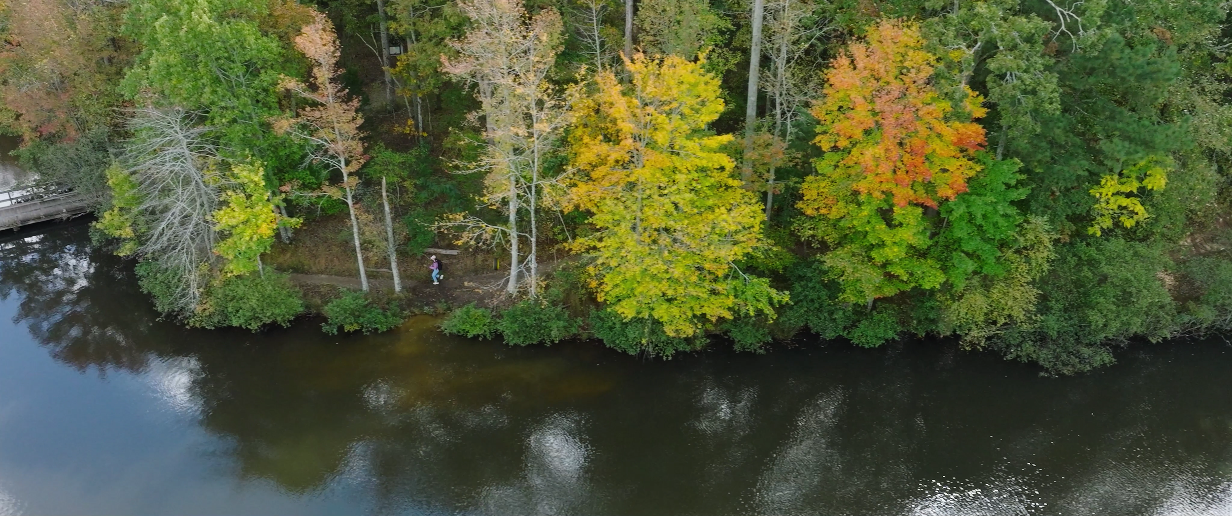 Aerial view of a riverbank with colorful autumn trees and two people walking on a path near the water.