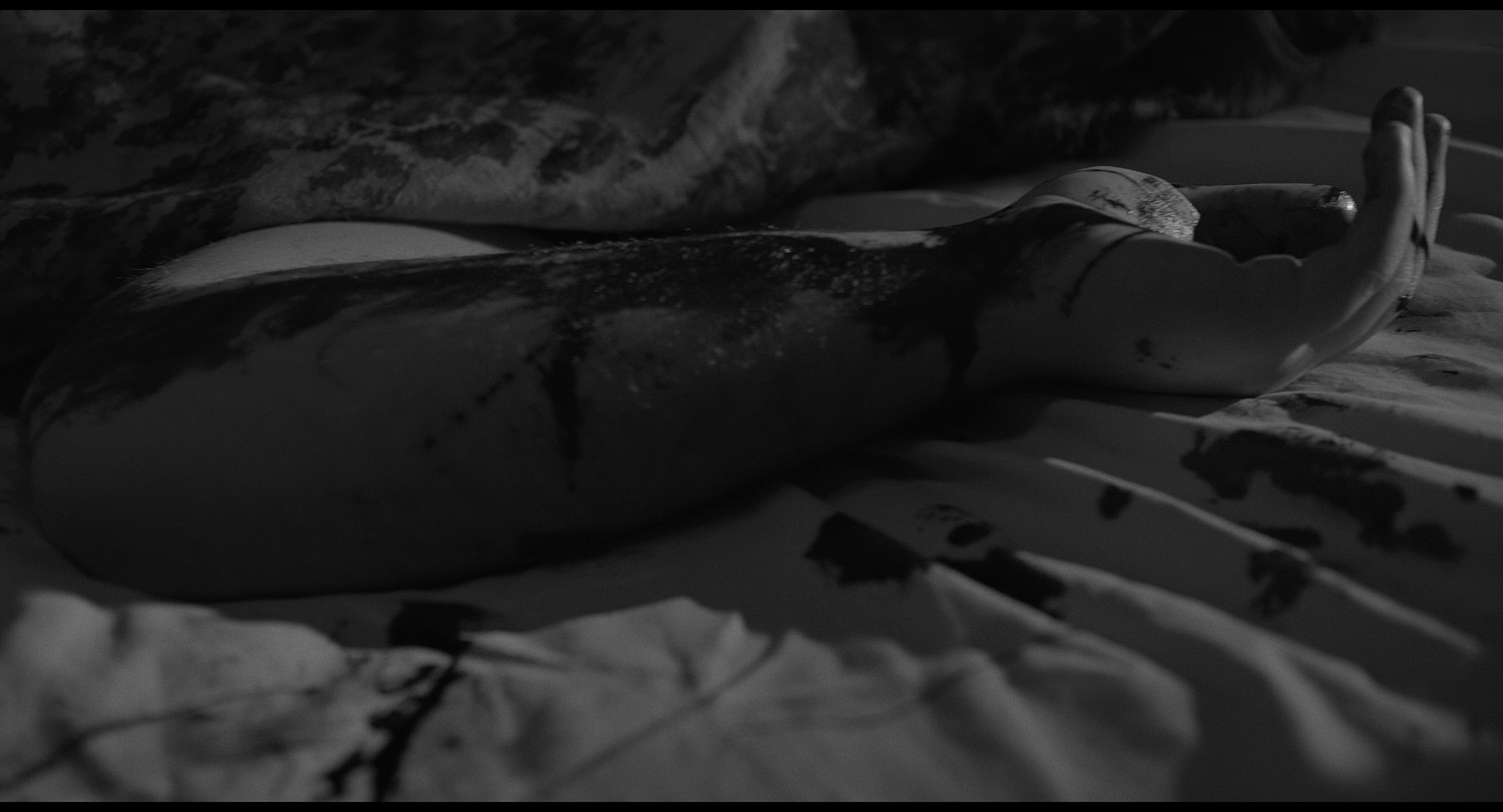 A close-up of a person's extended arm lying on a bed covered with a sheet with ink or paint splatters. The person is holding their hand up, with the palm facing upward.