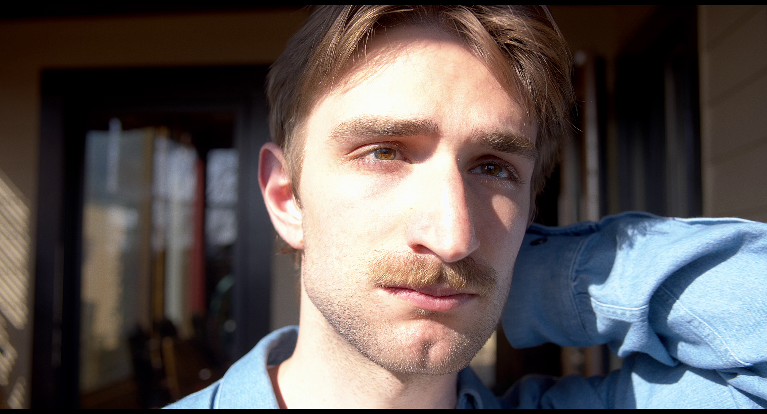 Close-up of a young man with brown hair, hazel eyes, and a mustache, wearing a blue shirt, outdoors during daytime.