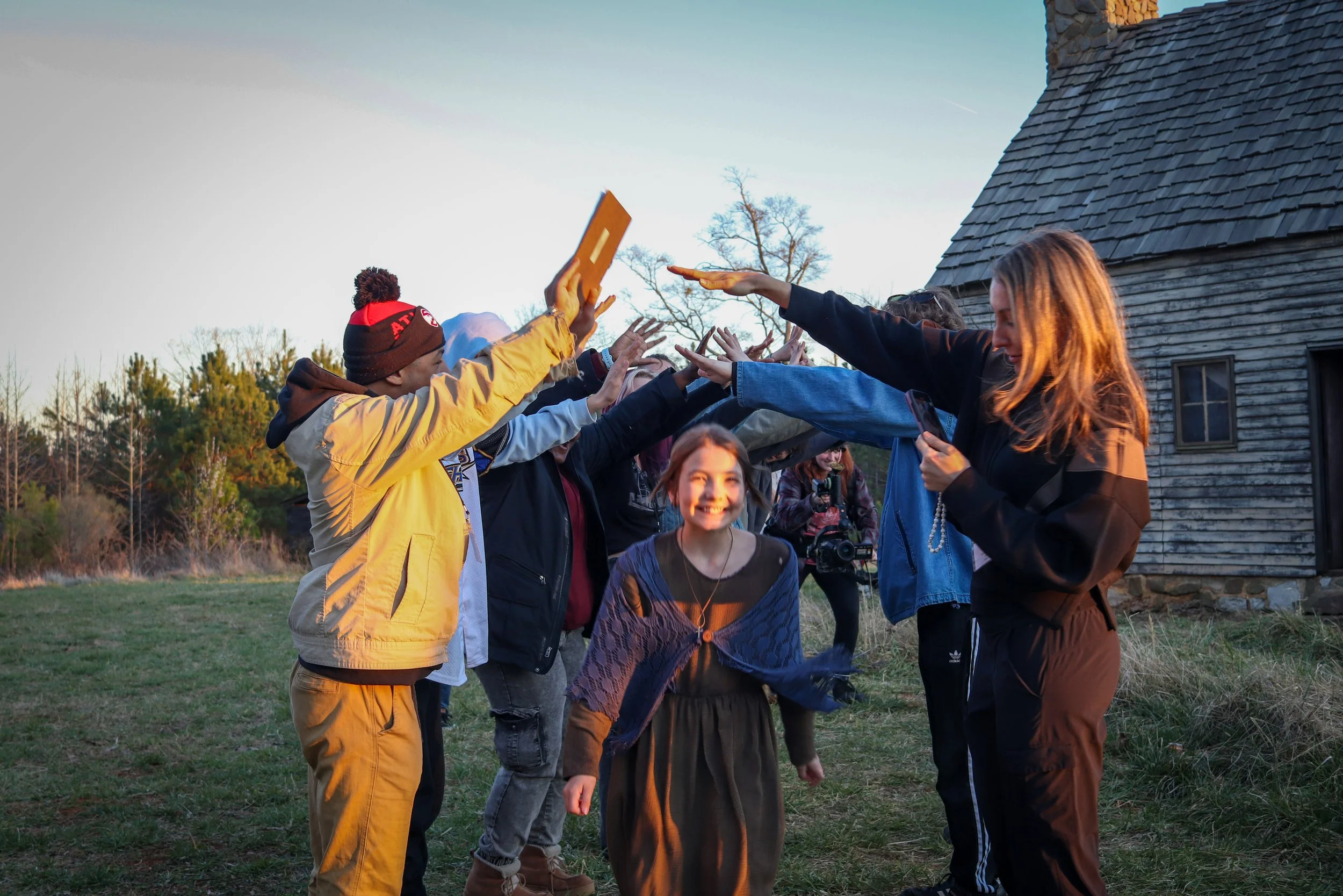 Group of people, including children and a woman, forming a tunnel with their arms for a smiling girl to walk through outdoors near a wooden building.
