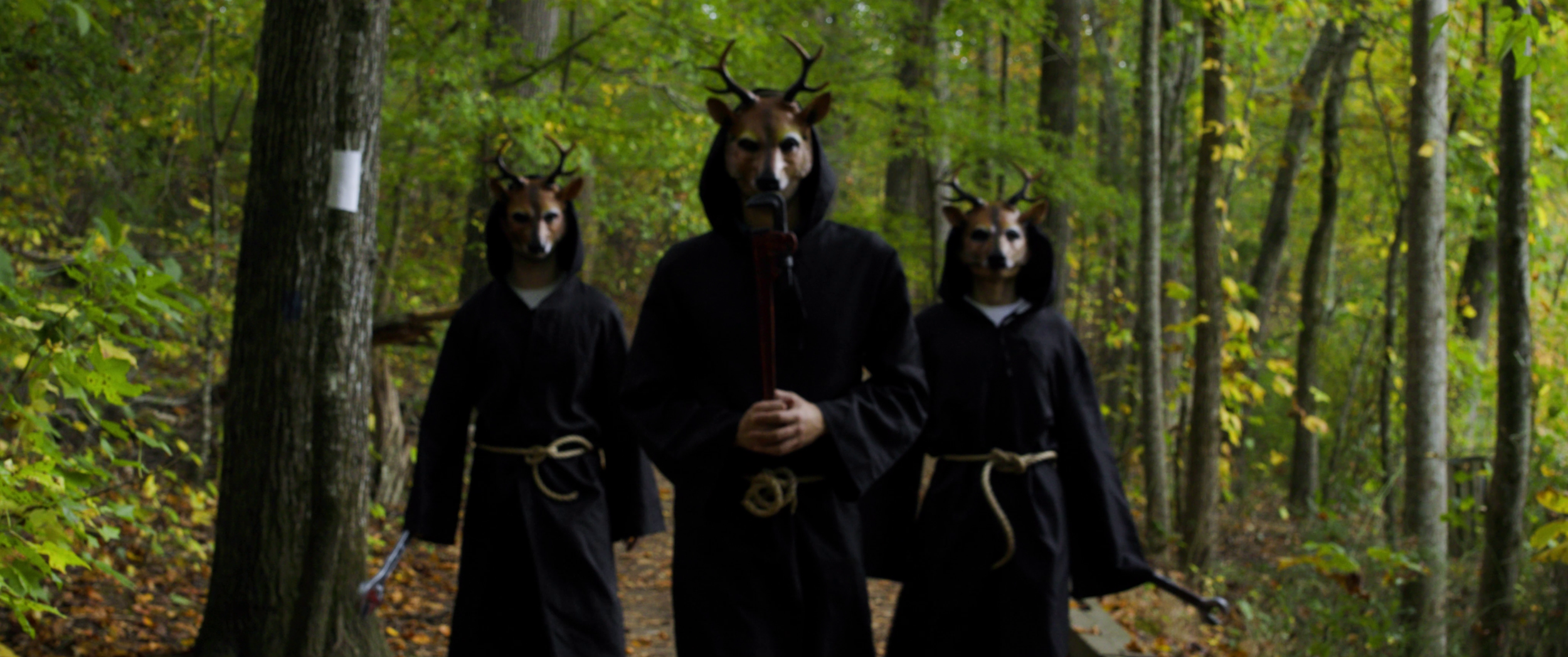 Three people in a forest, wearing black robes and animal masks with deer antlers, walking on a trail.