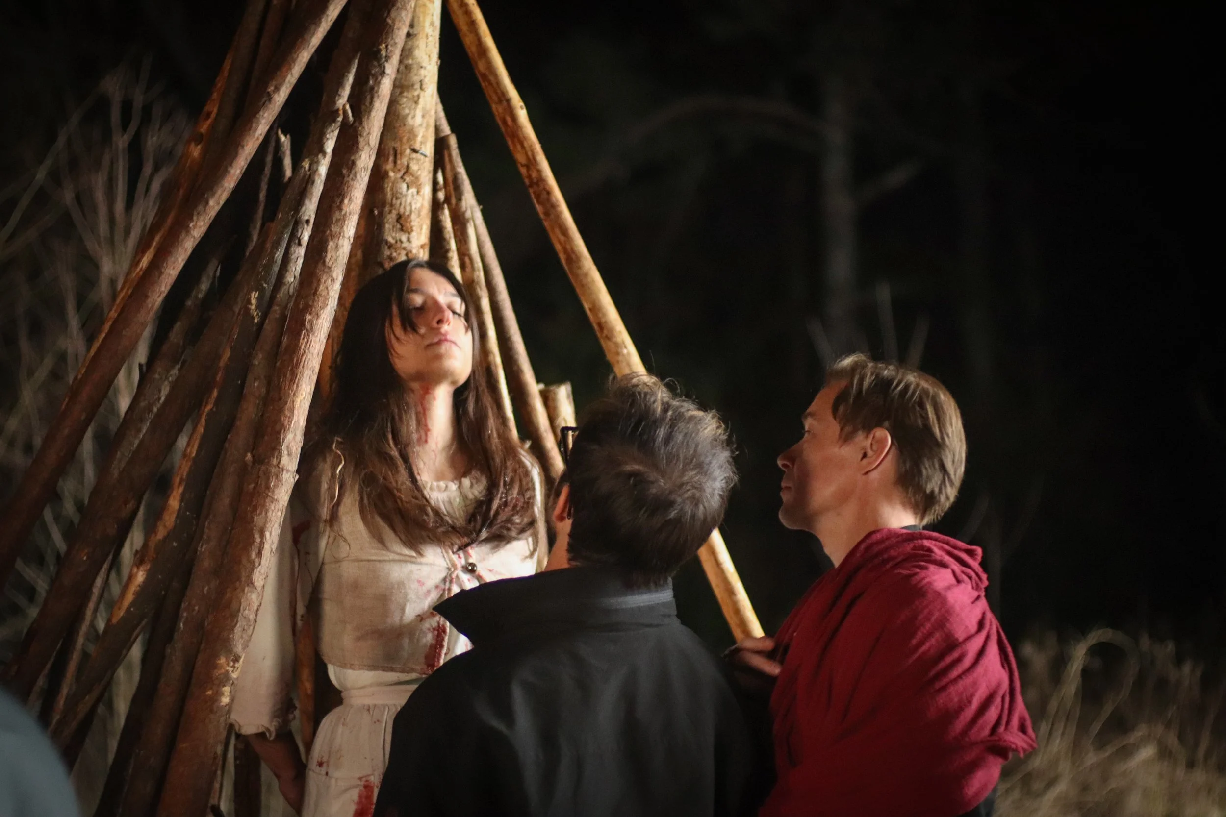 A woman with long brown hair and bloodstains on her clothes is tied to a wooden teepee structure, with her eyes closed. Two men, one in a black jacket and the other in a red cloak, are standing in front of her, appearing to be having a serious conver