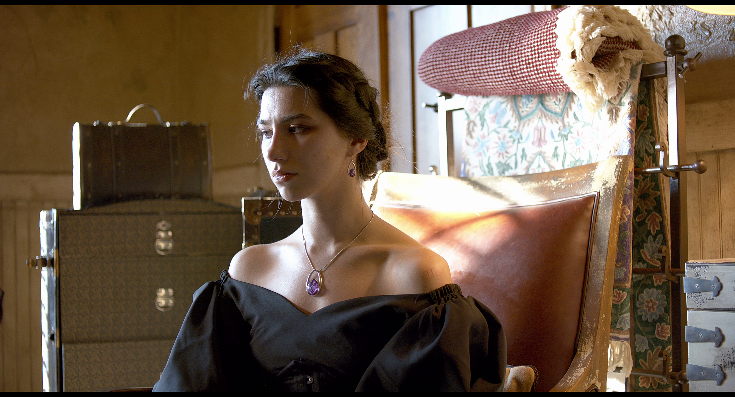 A young woman with short brown hair and purple jewelry sits on a wooden chair in a room with wooden walls. She wears a black off-the-shoulder top and looks contemplative. Behind her, there are stacked trunks and a roll of fabric or blanket on a rack.