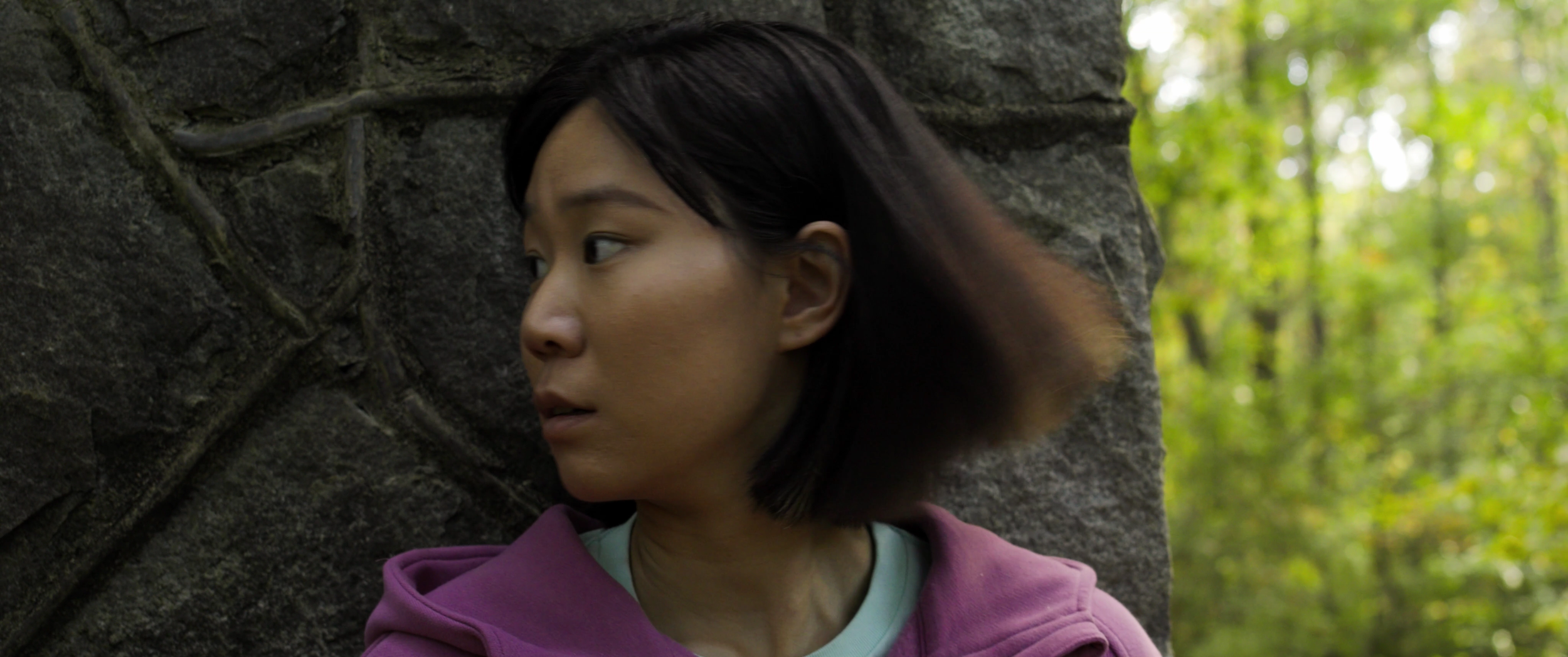 A woman with short black hair looking to her left outdoors next to a stone wall, with green trees in the background.