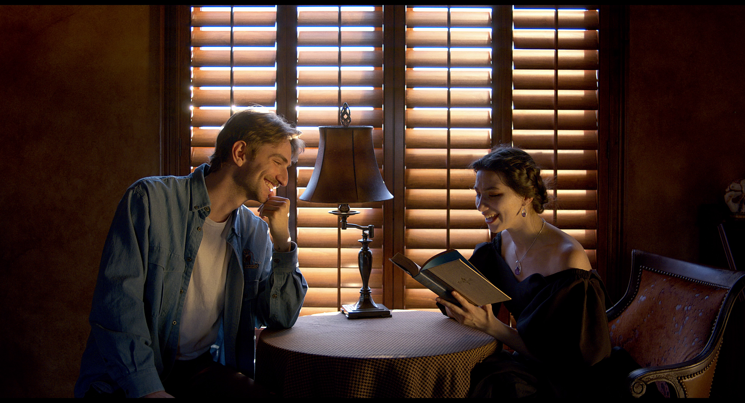A man and woman sitting at a small round table with a lamp, sharing a laugh while looking at a book, with window blinds behind them allowing sunlight to brighten the room.