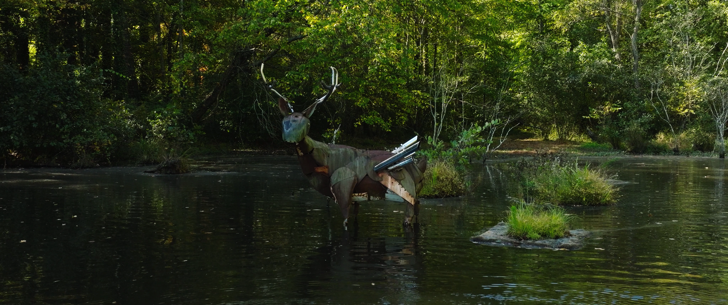A metal sculpture of a deer with large antlers, situated in a body of water surrounded by green trees and dense forest.