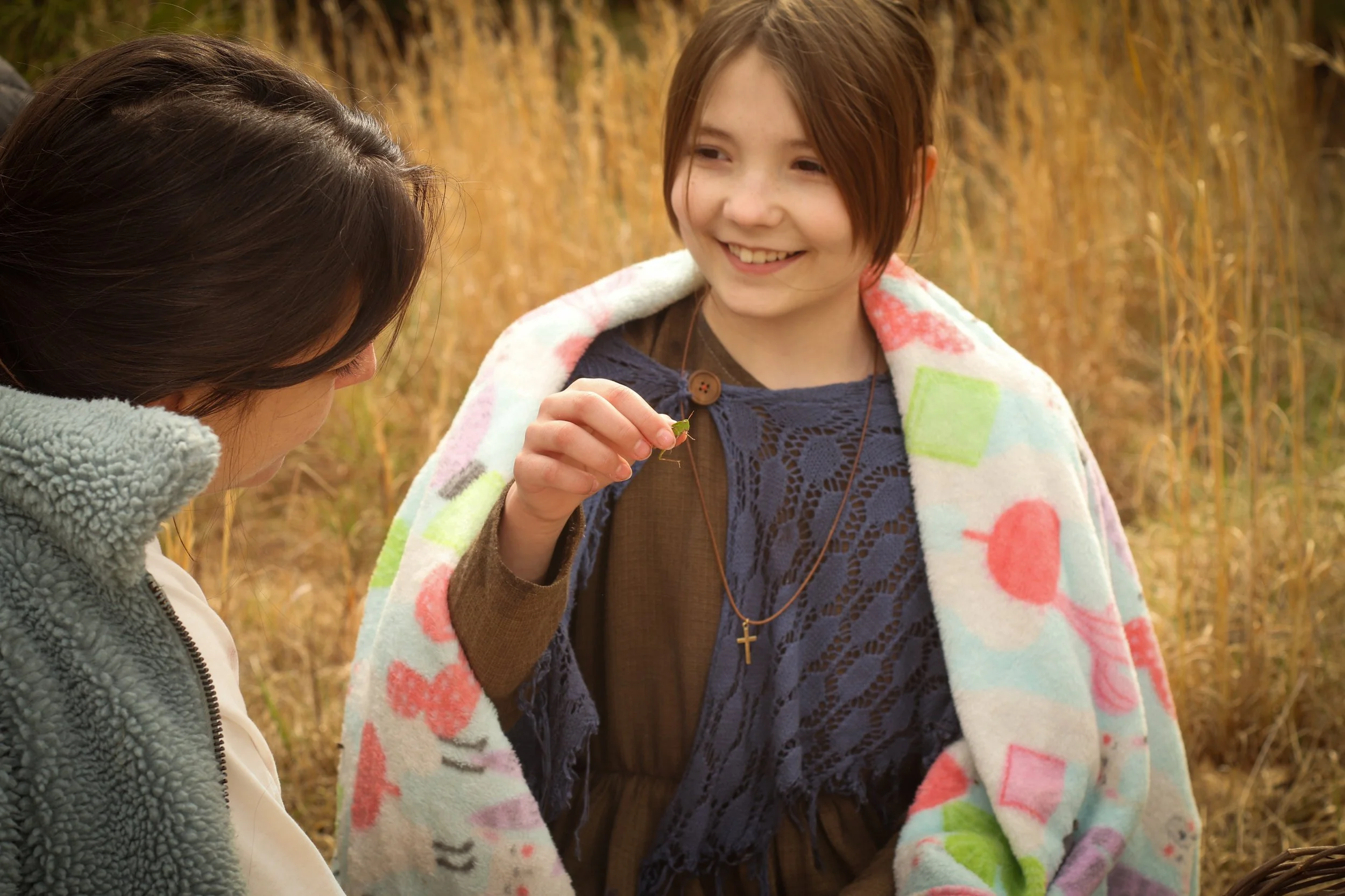 Two young girls outdoors, one holding a small green insect and smiling, wrapped in a colorful blanket, in a field of tall, golden grass.