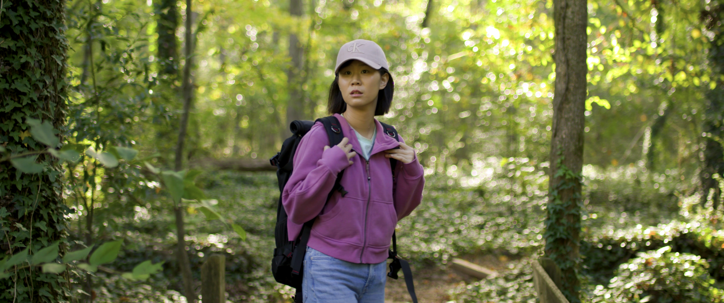 A young woman with shoulder-length black hair, wearing a light pink cap, purple jacket, and light blue jeans, stands in a lush forest with green trees and foliage, carrying a black backpack and looking to the side.