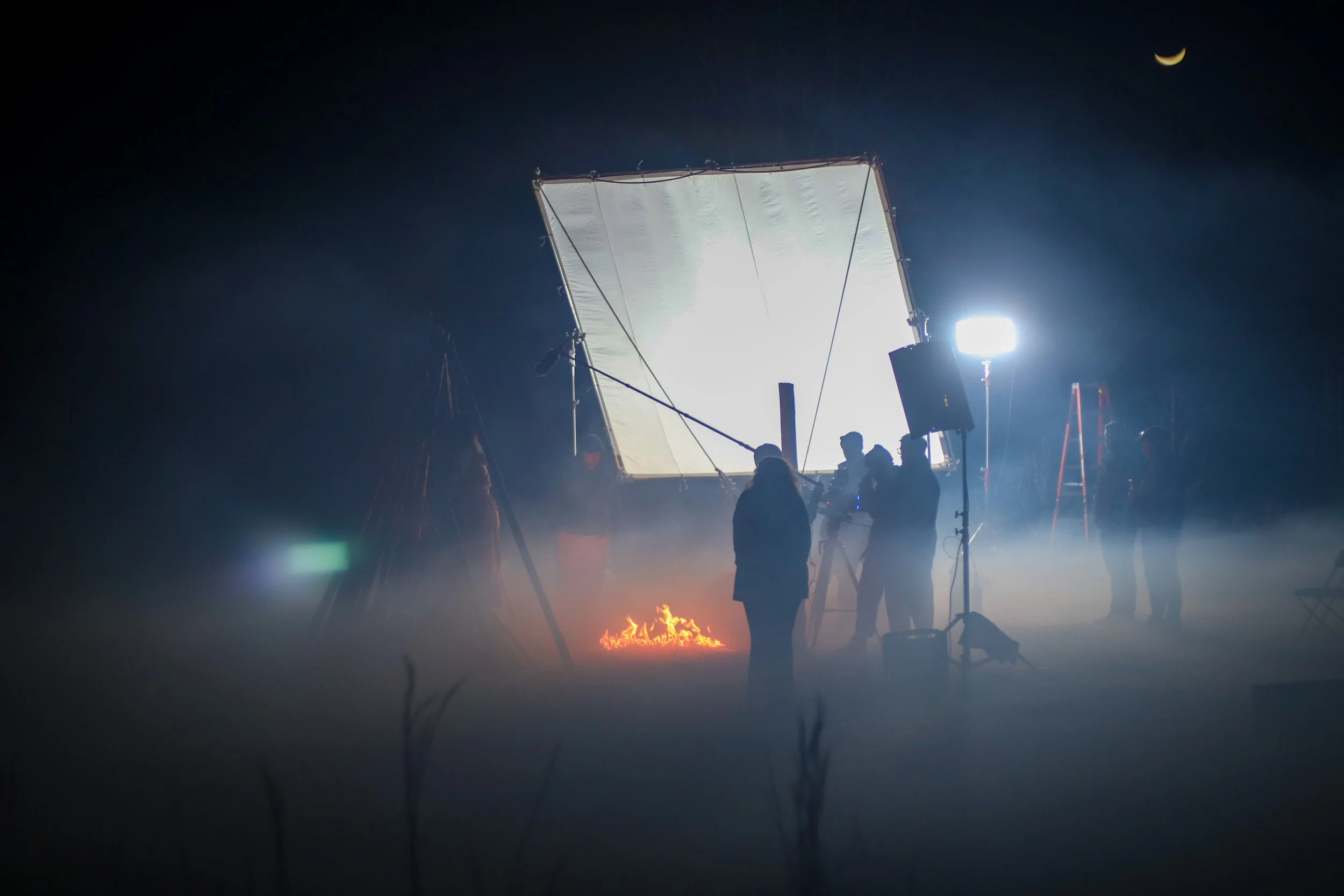 Film crew working outdoors at night with large lighting equipment, a fire on the ground, and a crescent moon in the sky.