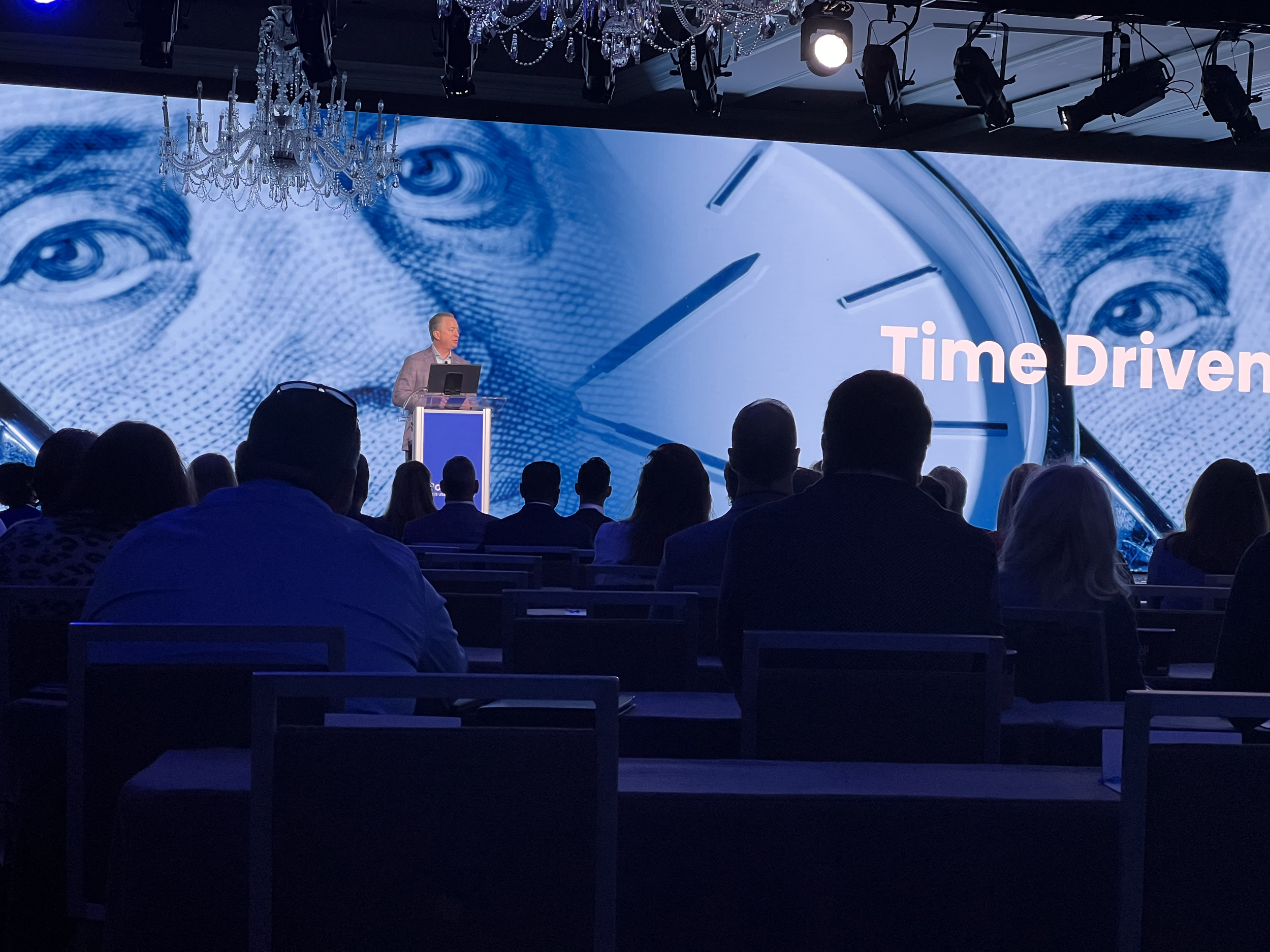 A man is giving a presentation on a stage with a large screen behind him showing a clock and the face of a person. The slide text reads 'Time Driven' and the audience is seated in a darkened room.