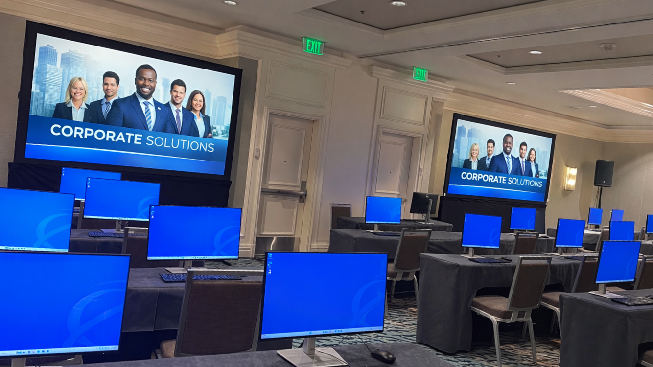 Large conference room set up for a presentation with multiple tables, each with a desktop computer and a chair. Two large screens display a corporate presentation titled 'Corporate Solutions' with images of diverse professionals in business attire. The room has a neutral color scheme with white walls, and there are exit signs above the doors.