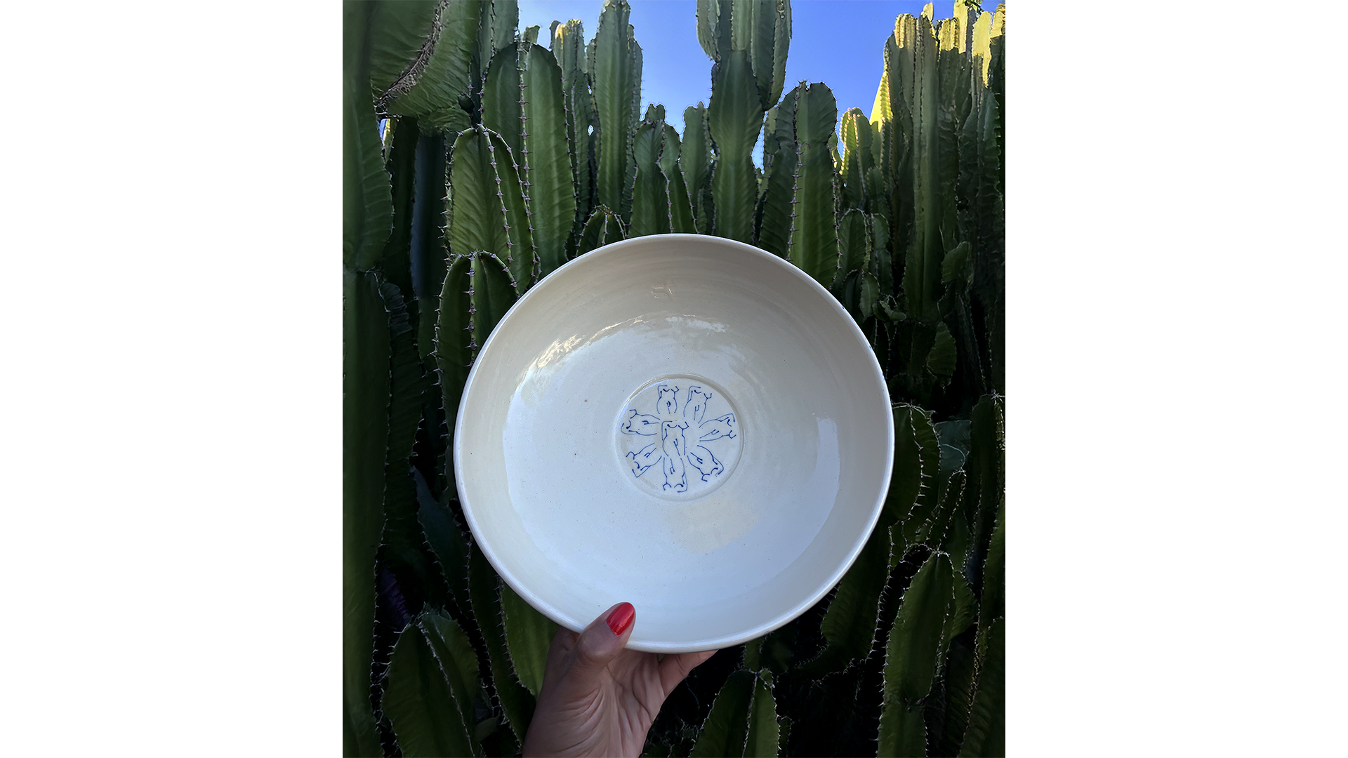 A person holding a white ceramic bowl with blue designs, against a background of tall green cacti and a clear blue sky.