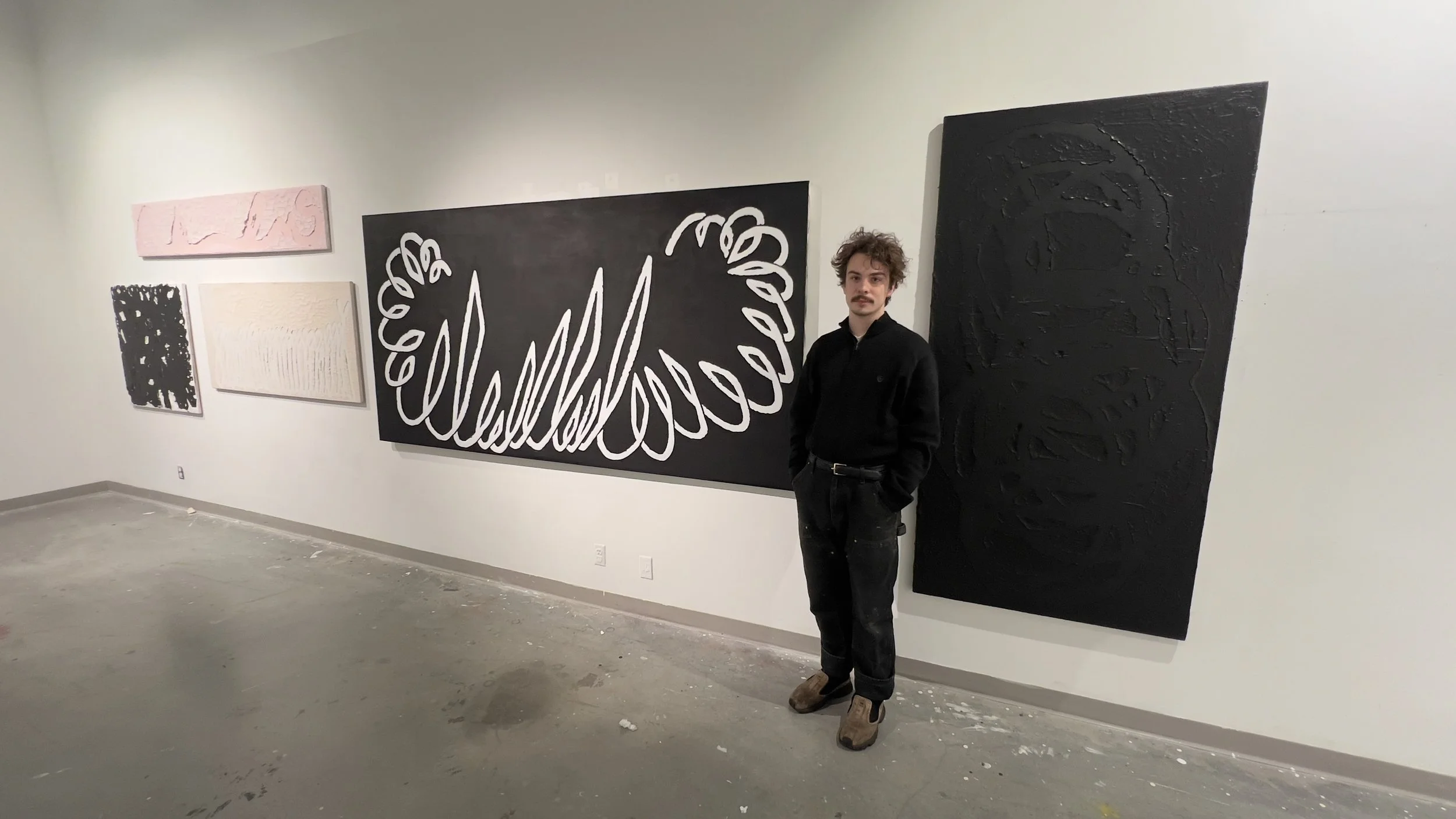 A man standing in an art gallery with six abstract paintings on the white wall behind him. The paintings are in various colors and styles, with the largest one being black and white with a looping design.