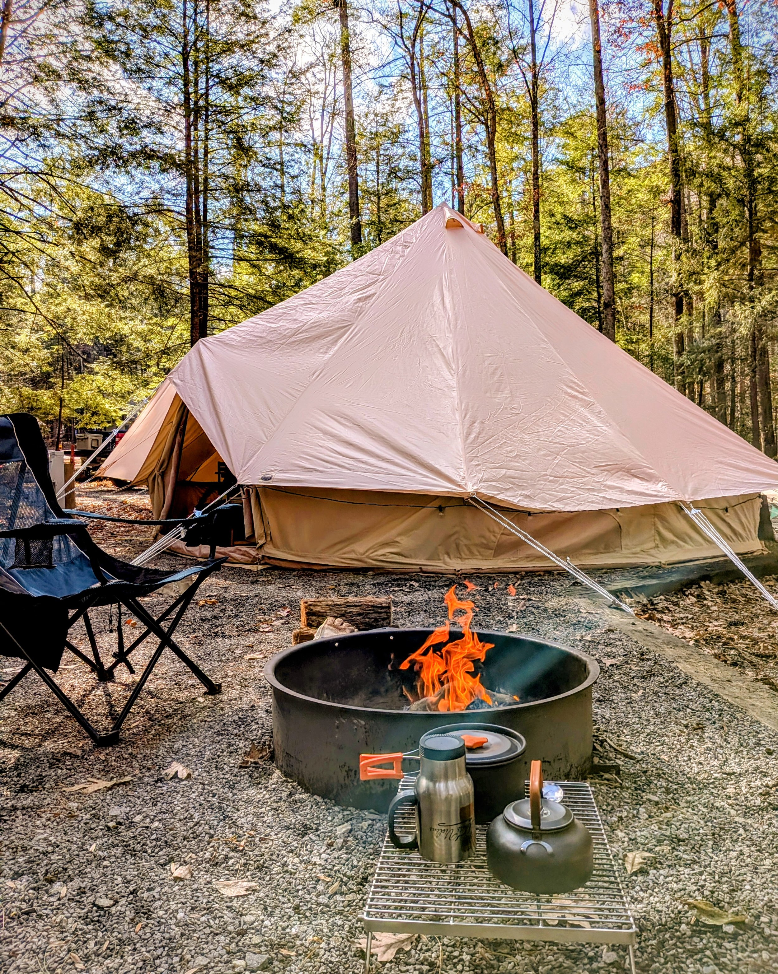 Glamping scene in a wooded area with a large beige bell tent, a camping chair, a fire pit with a small fire, and camping gear including a thermos and kettle. Trees in the background.