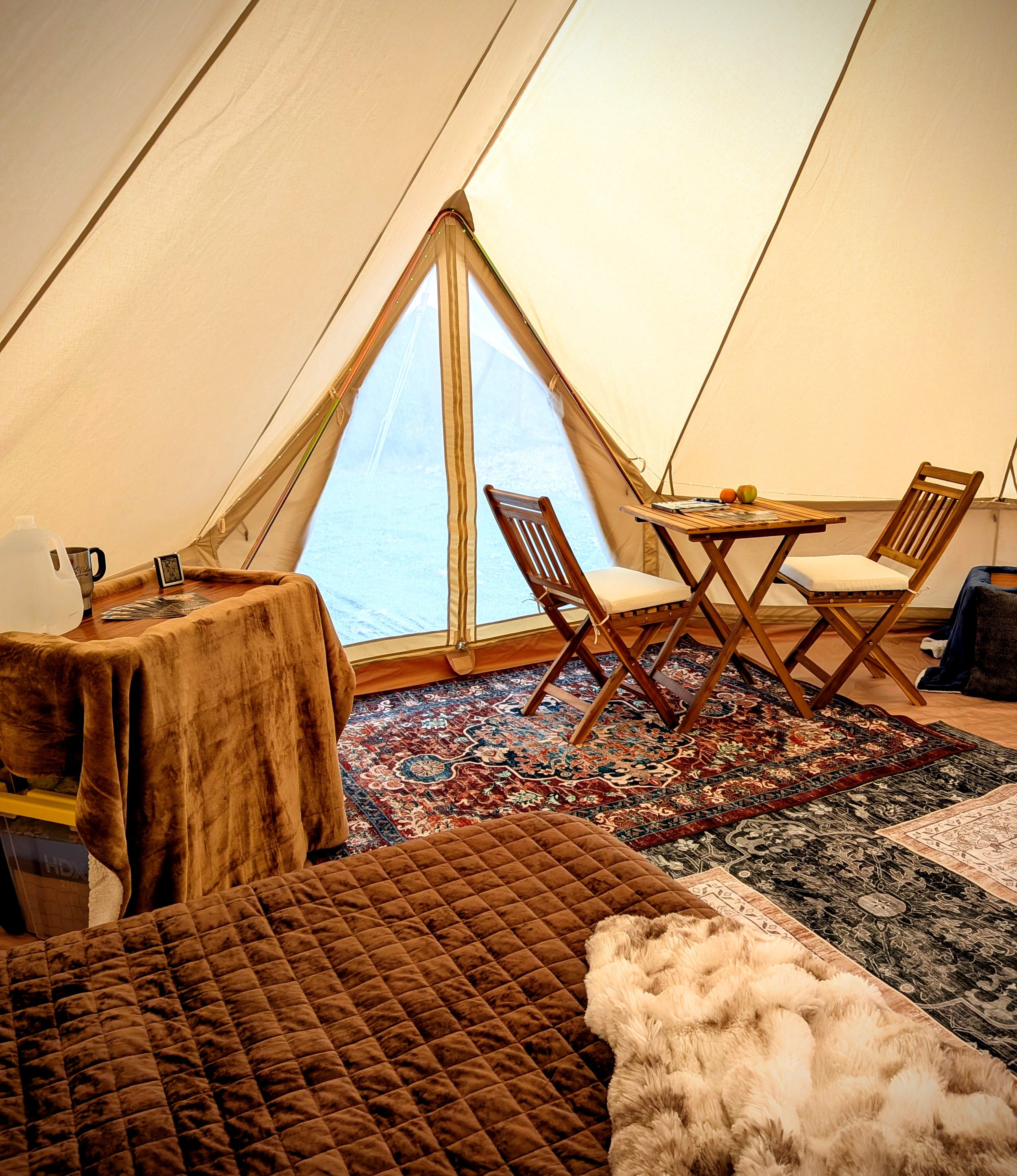 Inside a beige glamping bell tent with a sitting area, a small wooden table with two chairs, a large patterned rug, and various household items including a water jug, mug, and fur blanket.