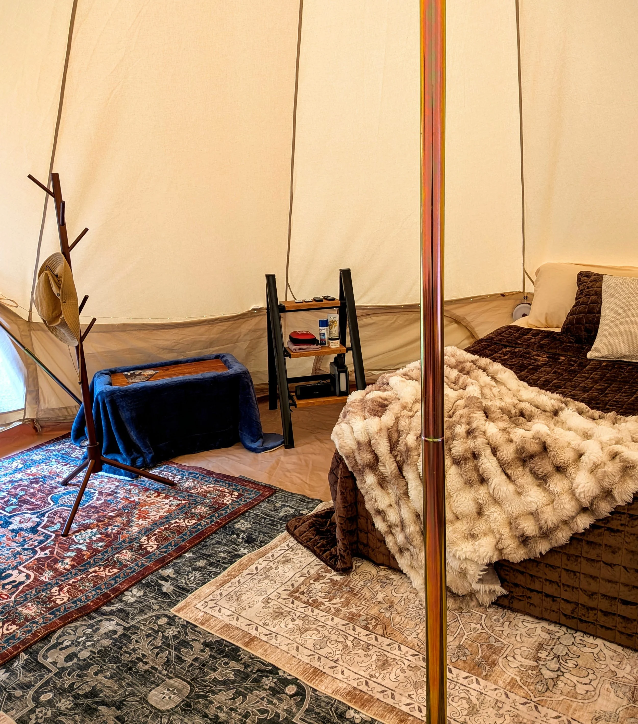Interior of a cozy glamping bell tent with a bed, patterned rugs, a small shelving unit with personal items, and a coat rack with a hat.