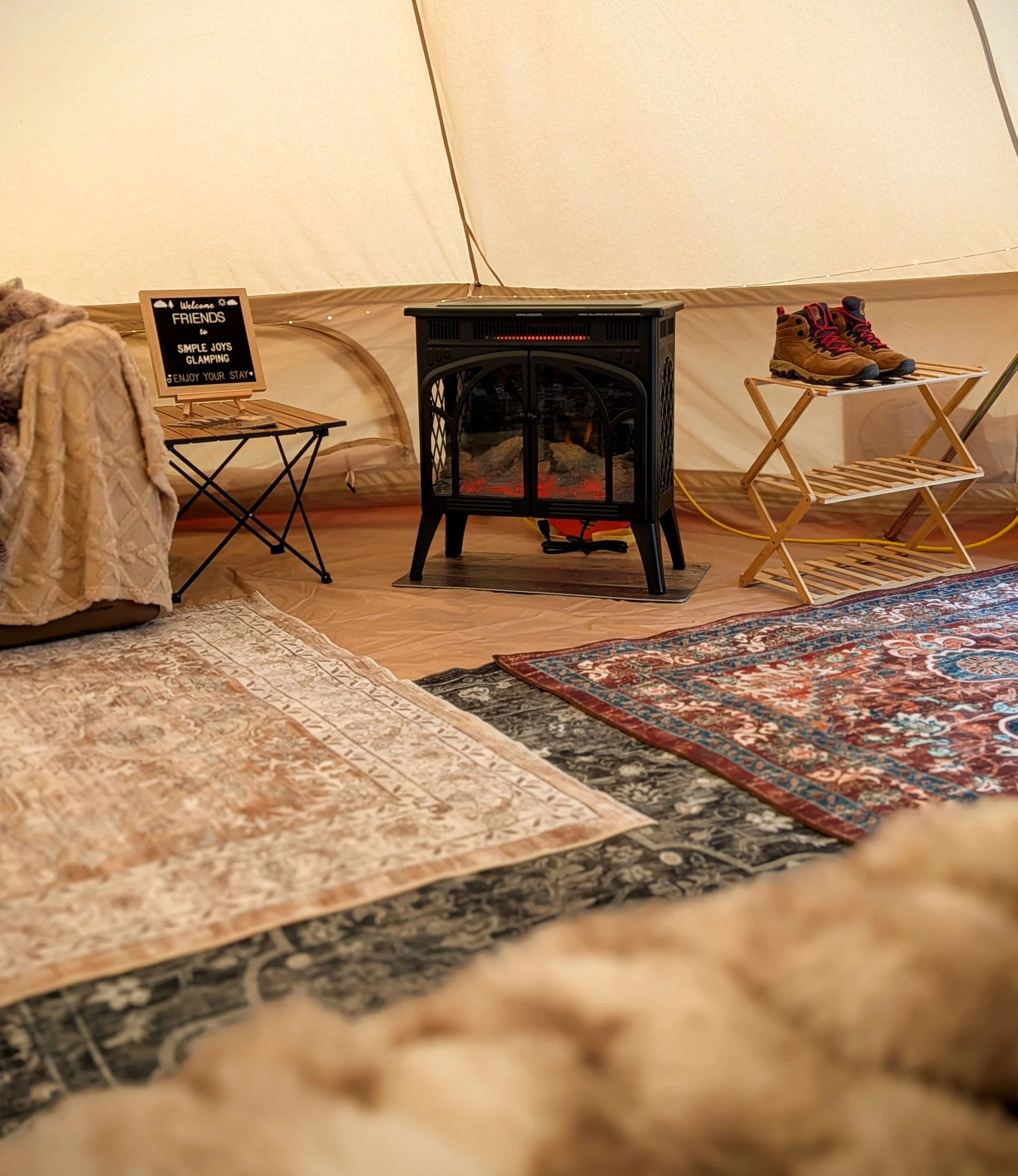 Interior of a cozy glamping bell tent with a small electric fireplace, a pair of hiking boots on a wooden rack, and decorative rugs on the floor.