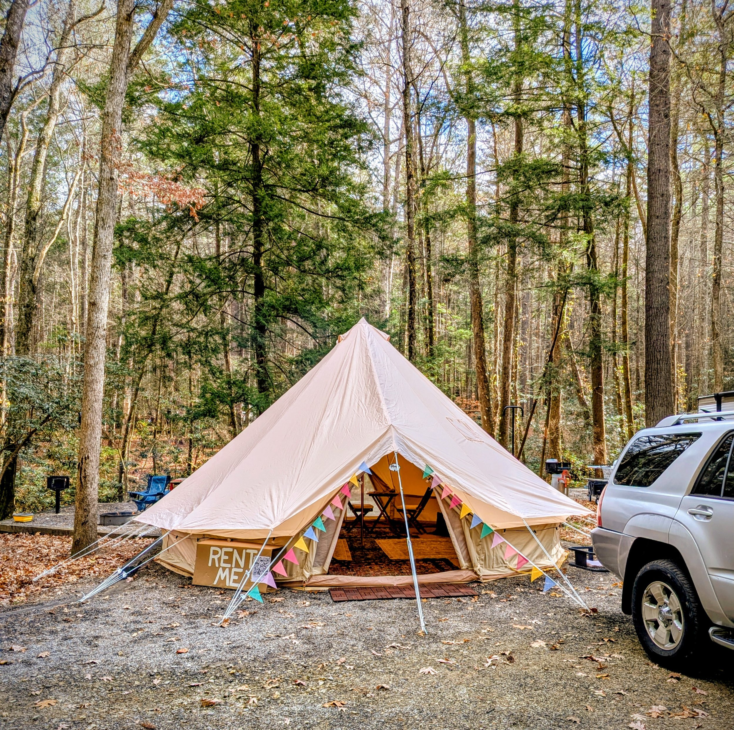 A large beige canvas glamping bell tent set up in a wooded area during daytime, decorated with colorful bunting, with a sign reading 'RENT ME' at the entrance.