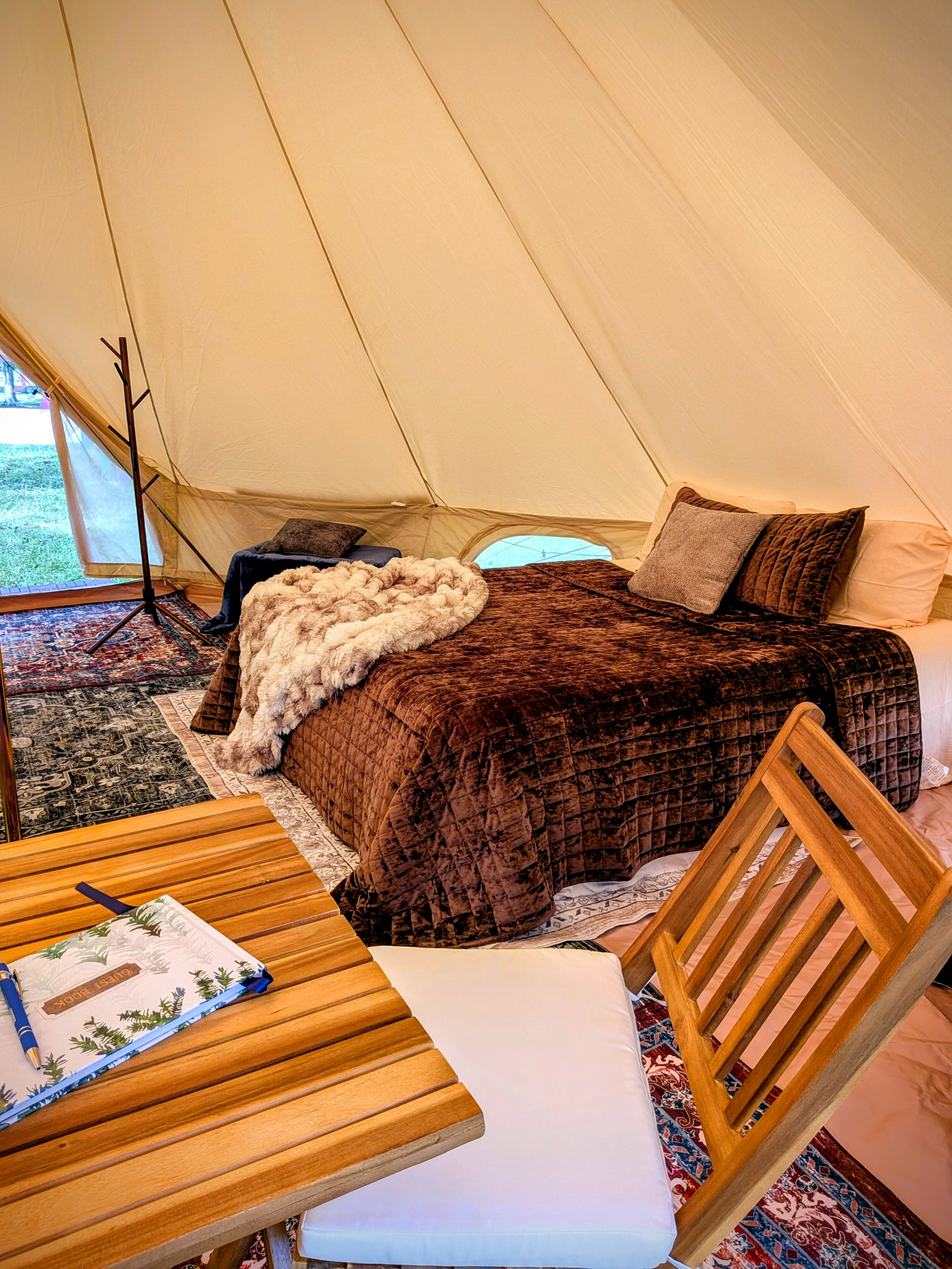 A cozy glamping bell tent interior with a bed covered in a brown quilt, accented by gray and brown pillows. There is a plush fur blanket on the bed, and a wooden table with a decorative notepad and pen nearby.
