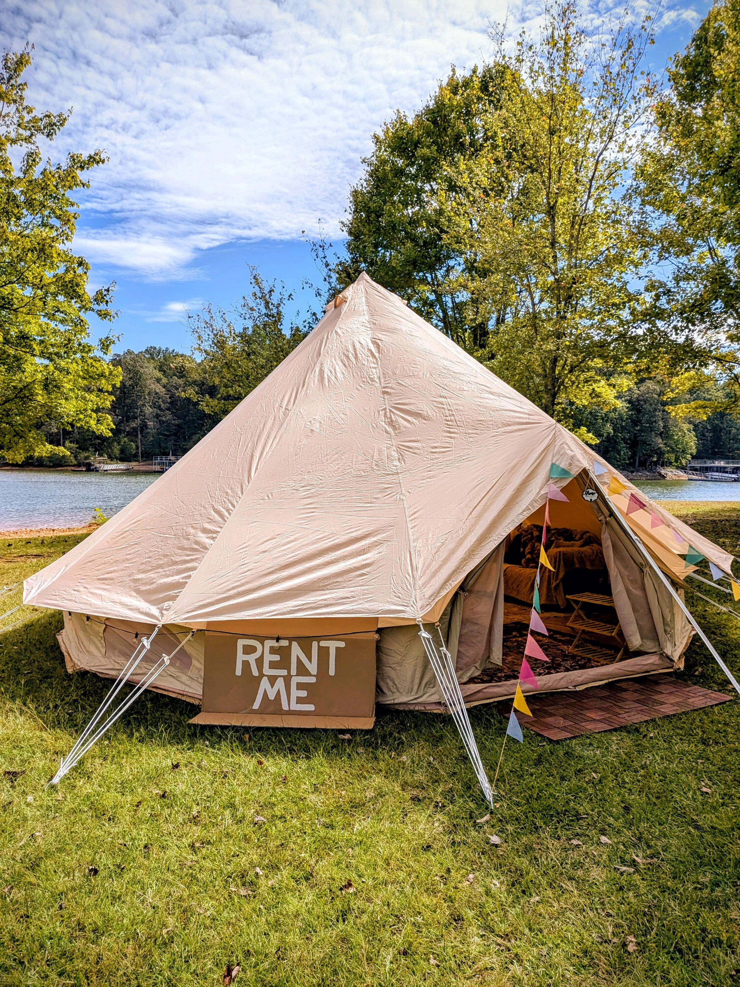 A large beige glamping tent pitched on a grassy area near Lake Lanier with trees in the background. The tent has a "RENT ME" sign and colorful pennant banners hanging on it.